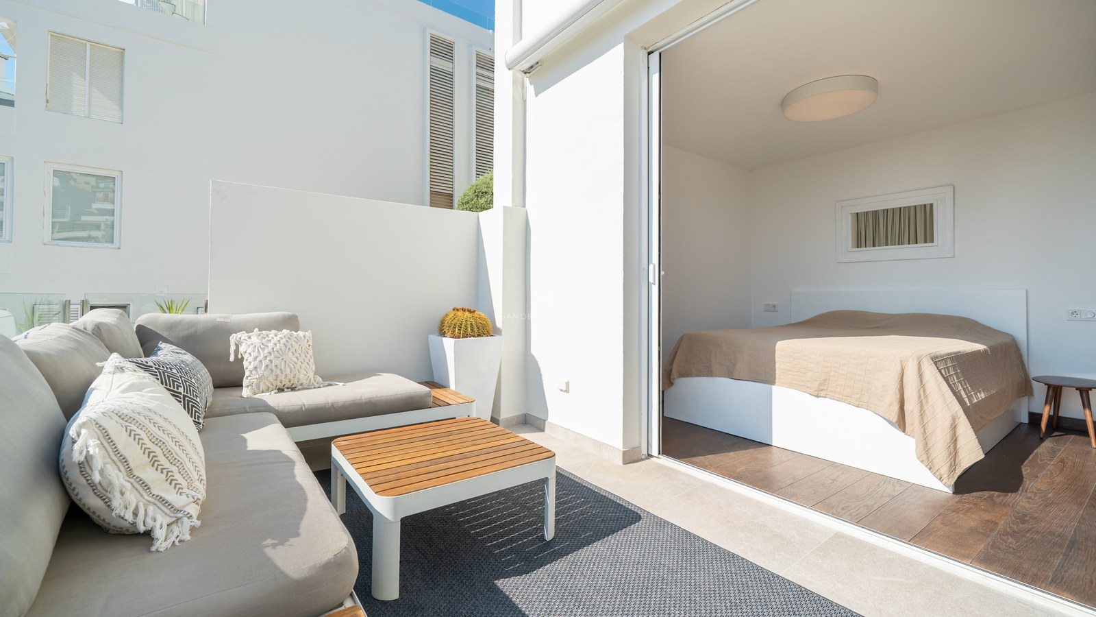 Dormitorio moderno abierto a una terraza soleada con sofás grises, mesa de madera y paredes blancas minimalistas.