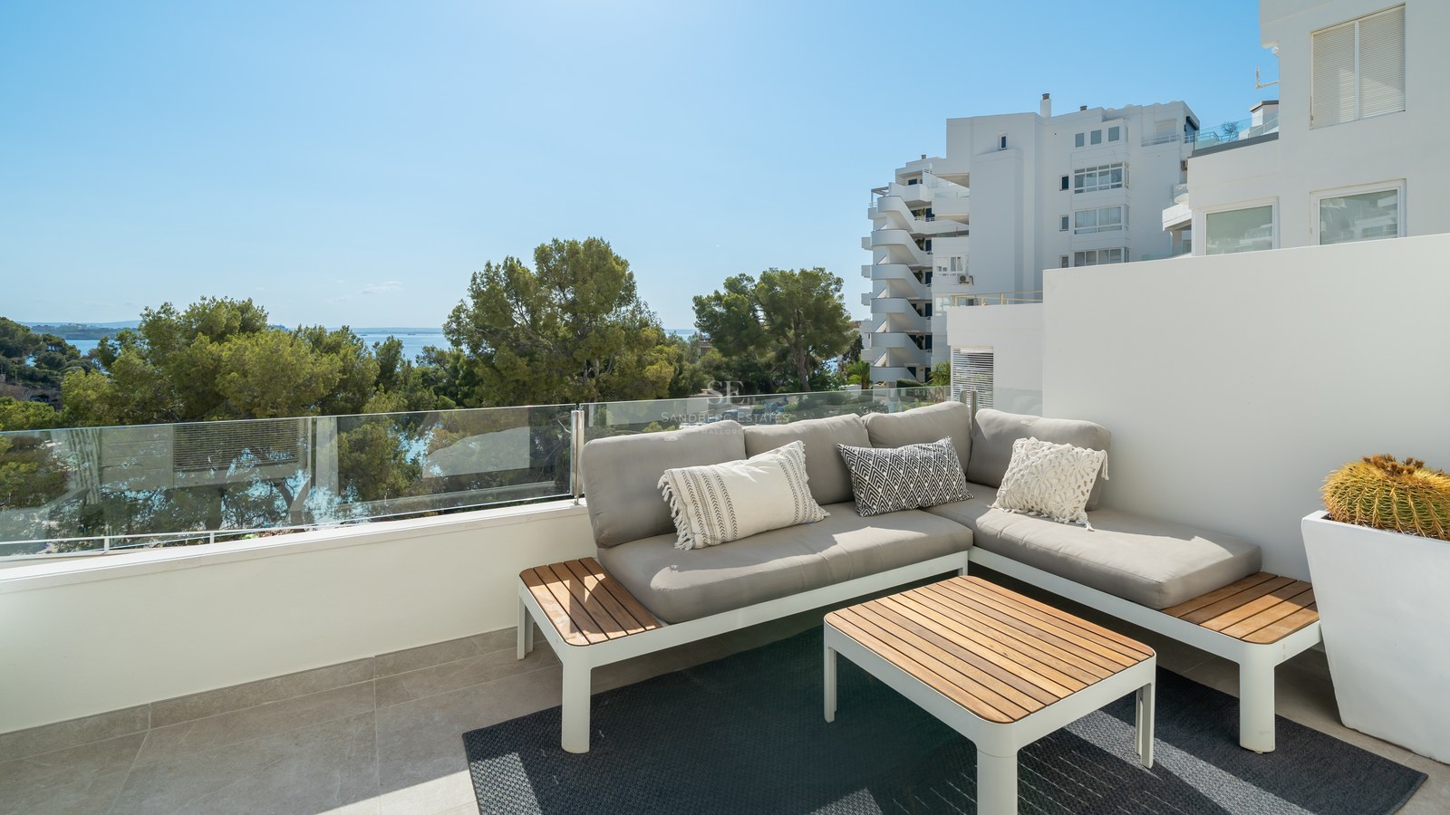 Terraza con sofá gris en L, mesa de madera y barandilla de cristal con vistas a pinos y al mar Mediterráneo.