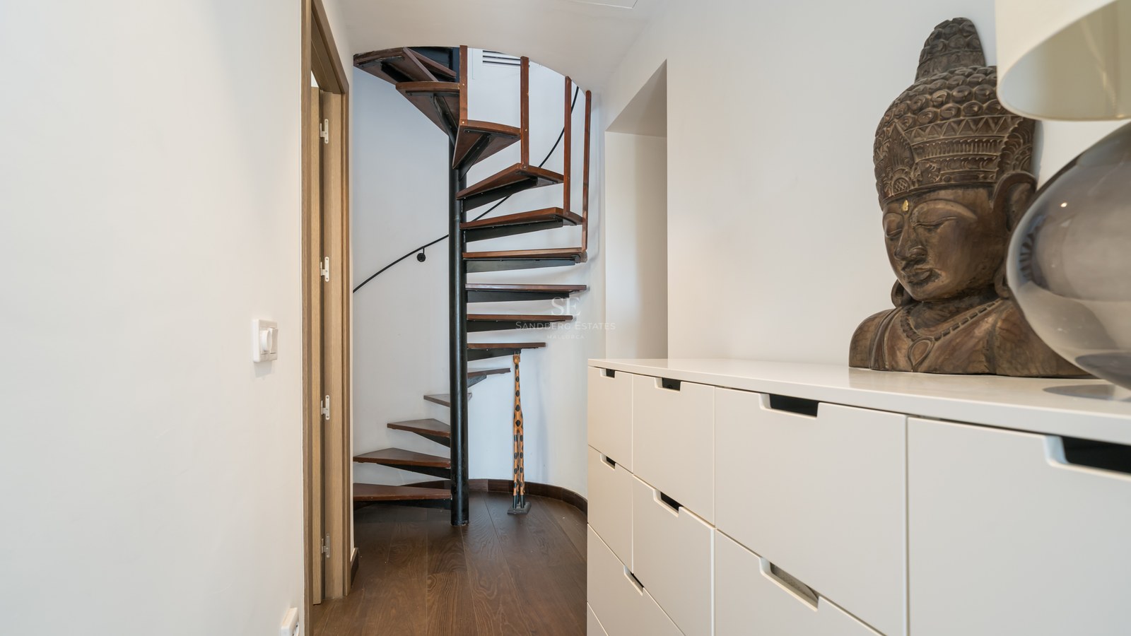 Pasillo moderno con escalera de caracol de madera oscura, mueble blanco y un busto de Buda decorativo.
