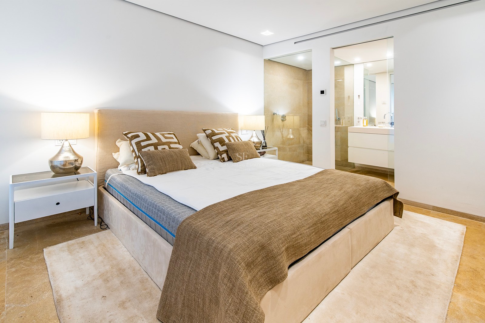 Chambre minimaliste avec lit beige, sol en pierre naturelle et salle de bains attenante vitrée.
