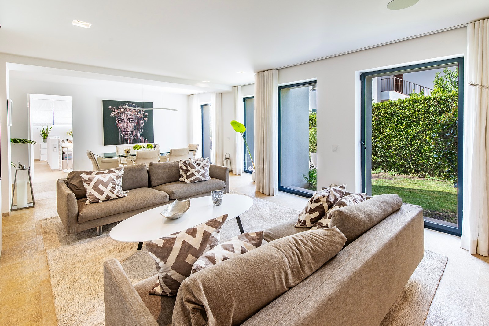 Salon lumineux avec canapés taupe et baies vitrées ouvrant sur un jardin privé bordé de haies.