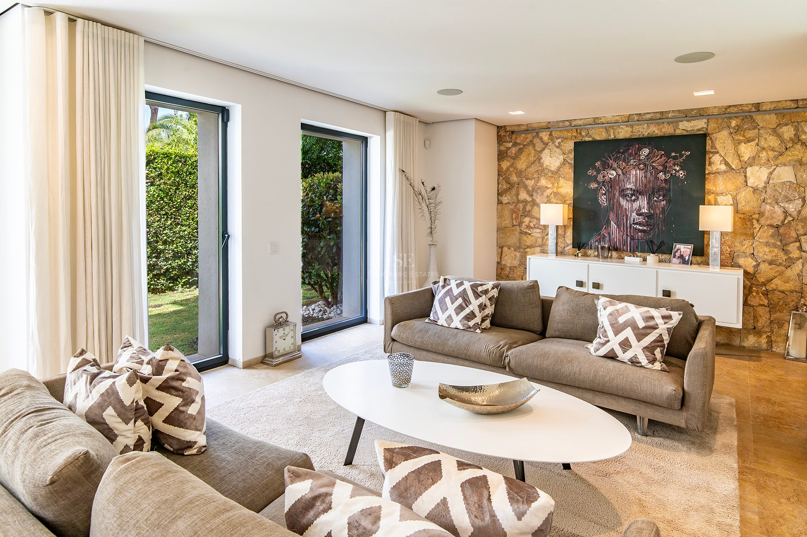 Salon moderne avec mur en pierre naturelle, canapés taupe et portes vitrées s'ouvrant sur un jardin verdoyant.