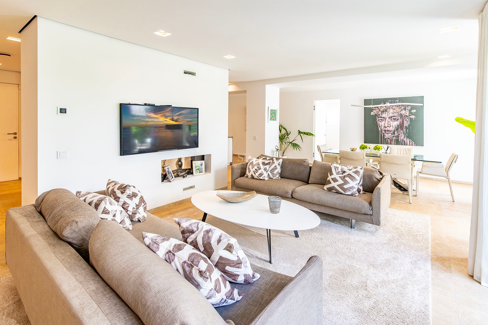 Vaste salon moderne avec canapés taupe, table basse ovale blanche et salle à manger avec œuvre d'art en arrière-plan.