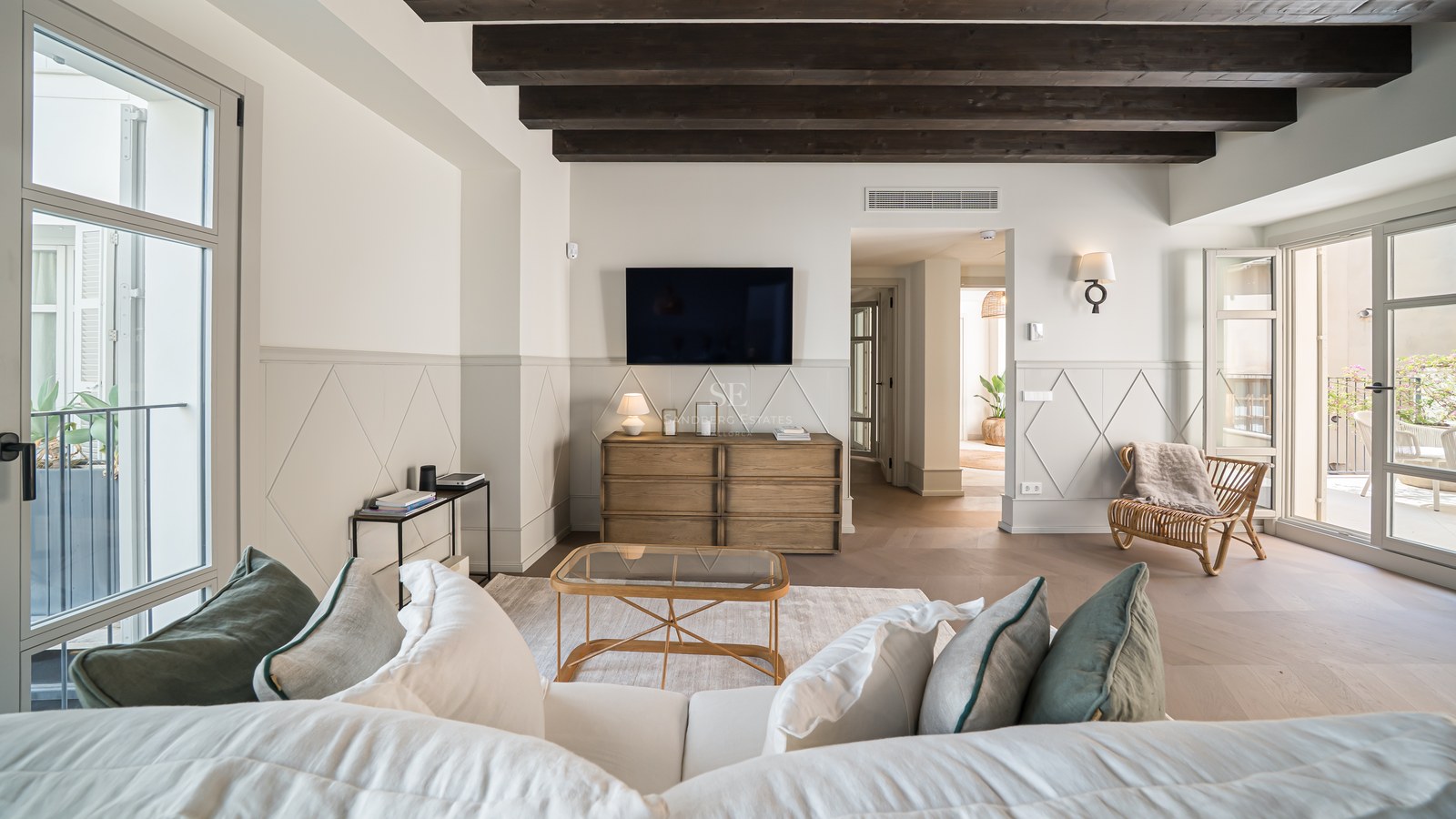 Bright living room featuring dark exposed ceiling beams, decorative wall paneling, and large windows leading to a balcony.