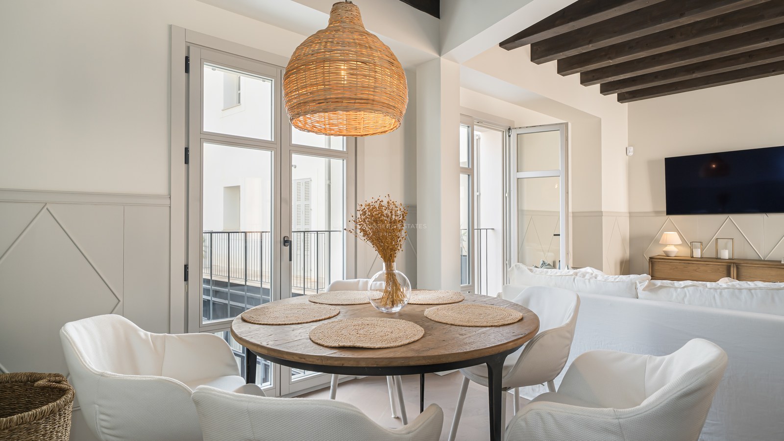 Modern round wooden dining table with white chairs, wicker pendant light, and dark exposed ceiling beams.