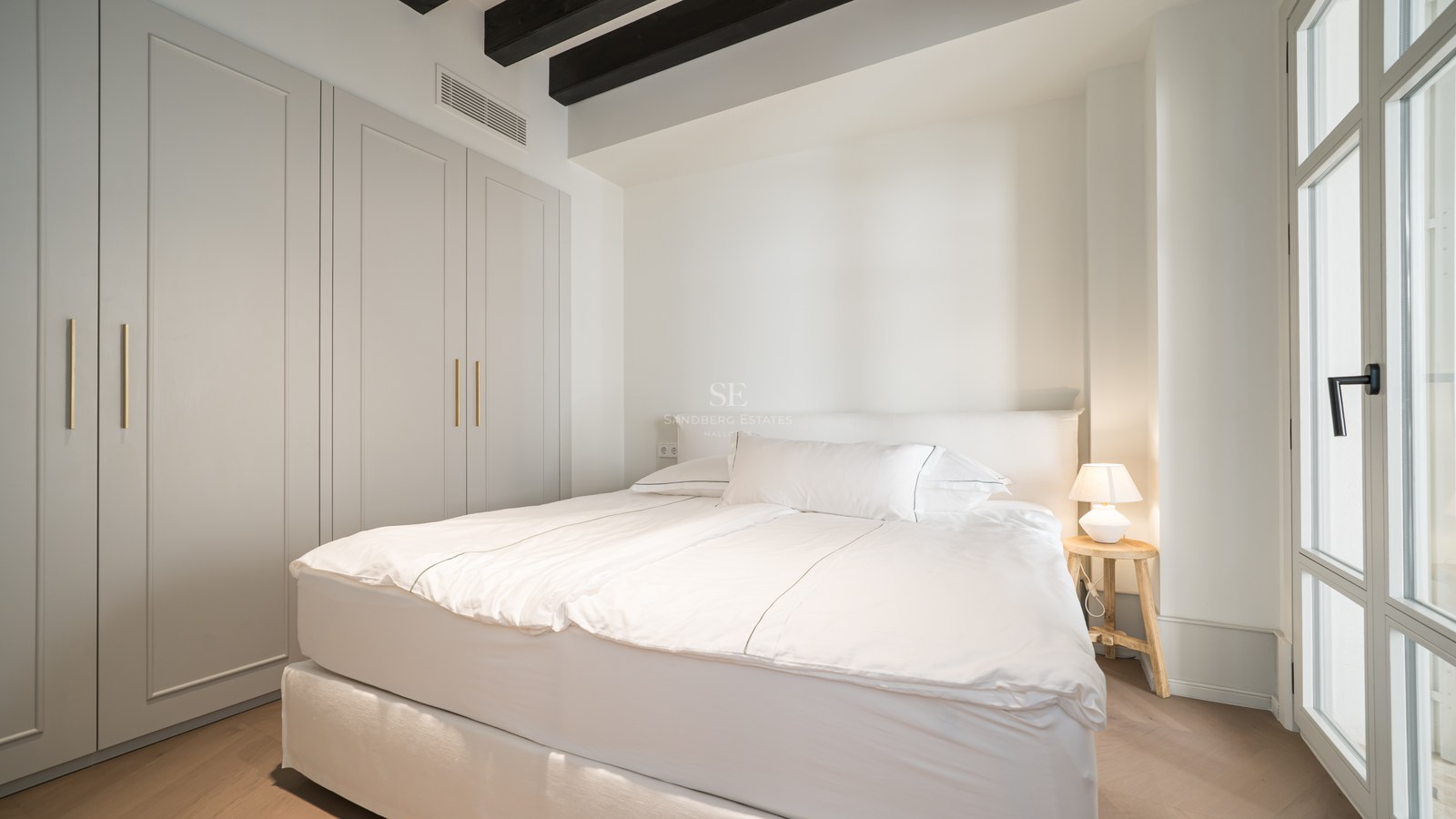Bright bedroom with white linens, dark wood ceiling beams, built-in wardrobes, and light oak flooring.