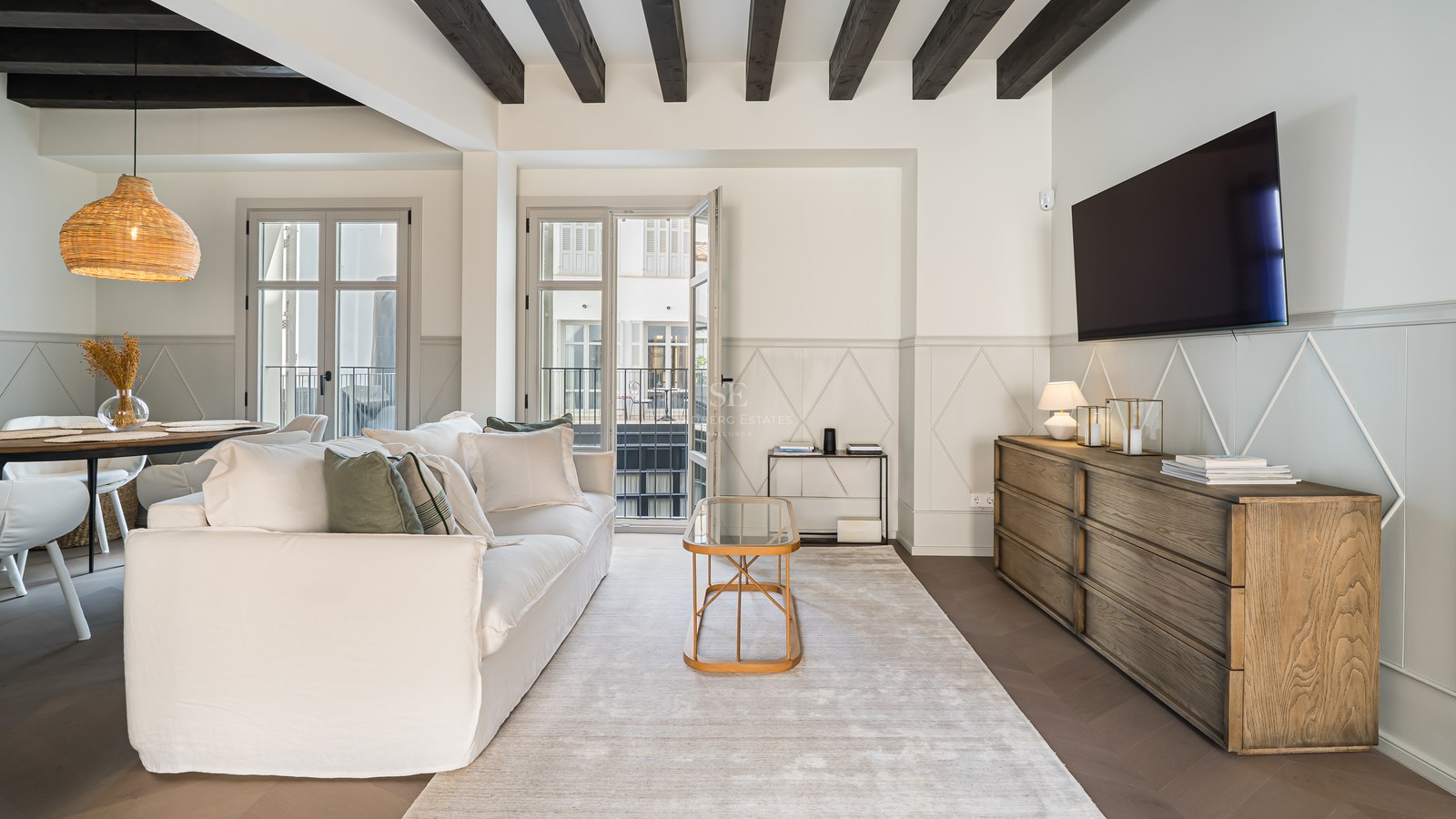 Bright living room featuring a white sofa, dark wooden ceiling beams, a wooden sideboard, and French balcony doors.