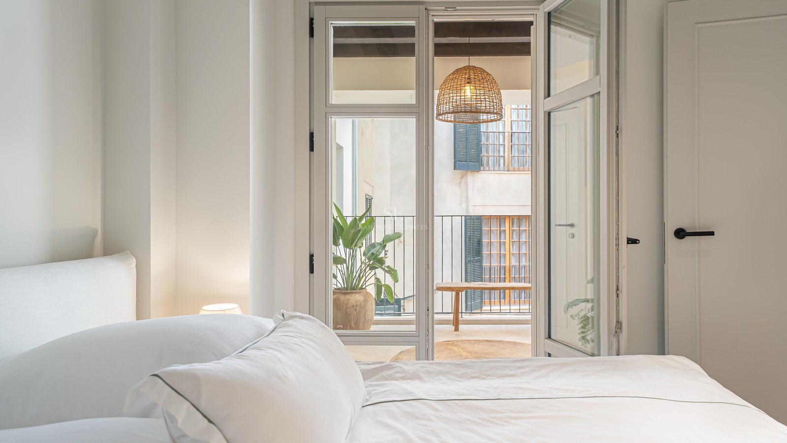 A bright bedroom with white linens looking through open glass doors to a balcony with a woven lamp and plants.