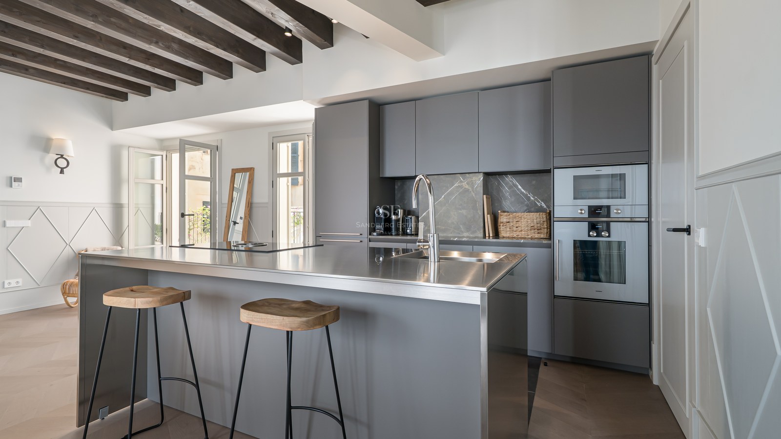 Modern kitchen featuring grey cabinetry, a stainless steel island with bar stools, and exposed wooden ceiling beams.