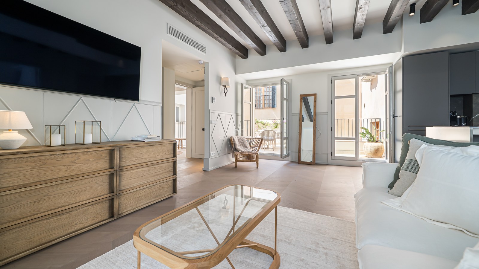 Bright contemporary living room with dark wooden ceiling beams, oak furniture, and doors opening to a balcony.