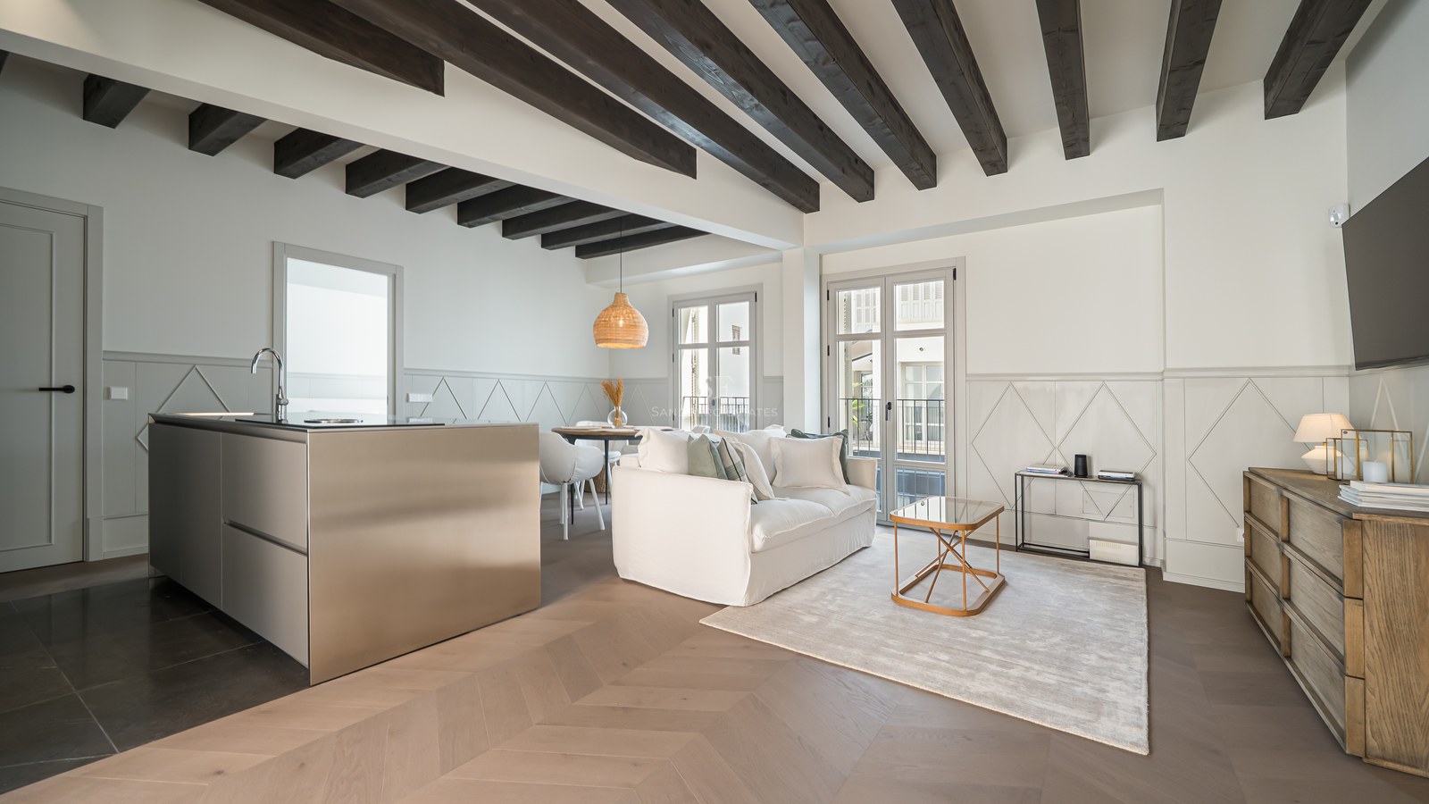 Modern living room with dark wood ceiling beams, chevron floors, and a sleek stainless steel kitchen island.