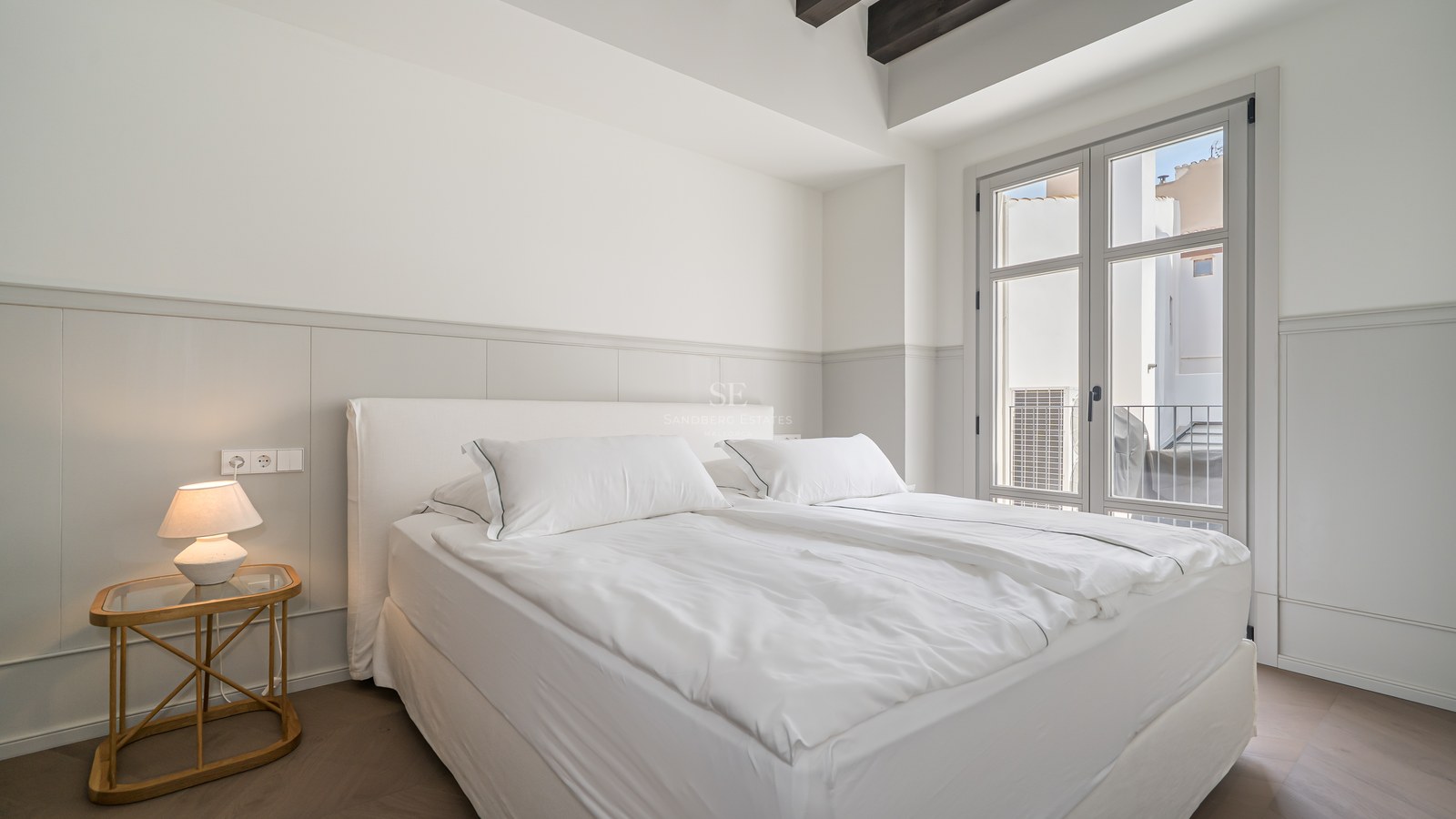 Bright bedroom featuring white linens, dark wooden ceiling beams, and a French window opening to a Juliet balcony.