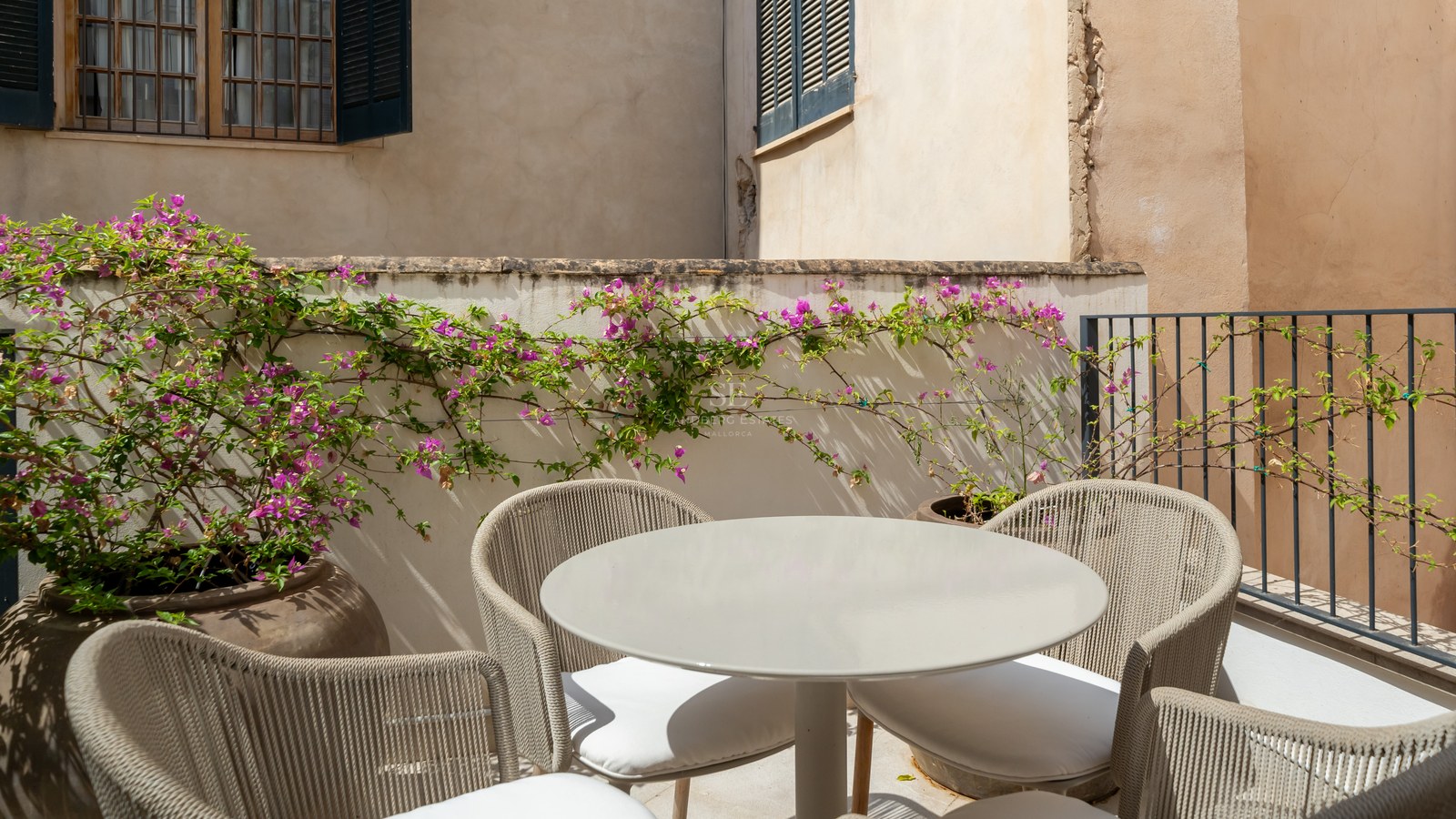 A round grey outdoor table and four woven chairs on a sun-drenched terrace with vibrant pink bougainvillea.