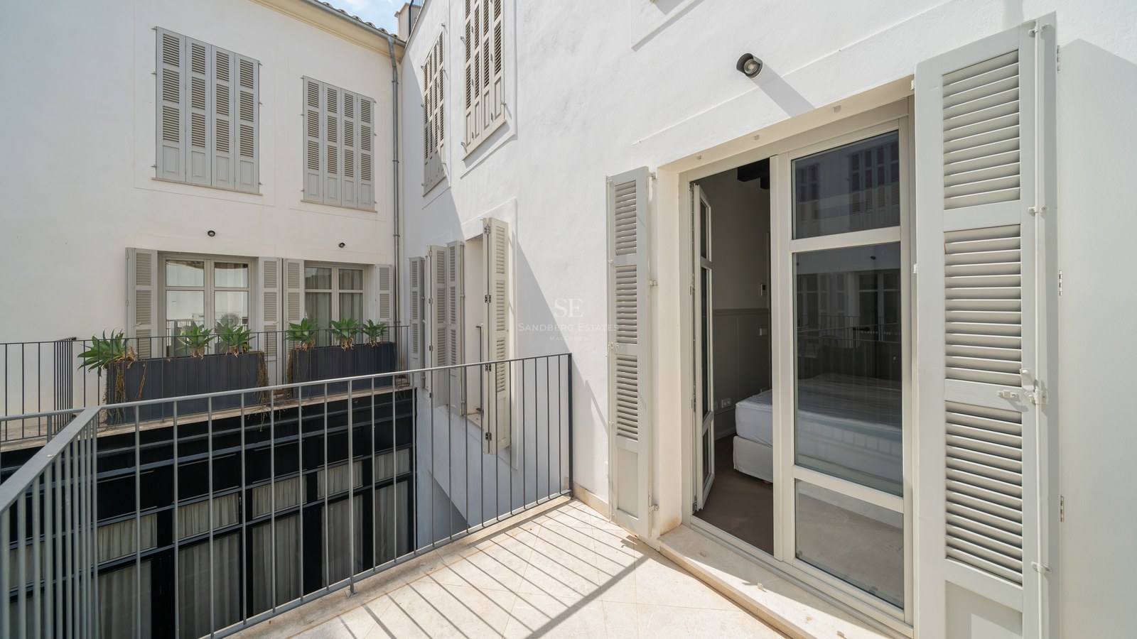Private terrace with light stone flooring, black iron railings, and grey wooden shutters on a clean white facade.