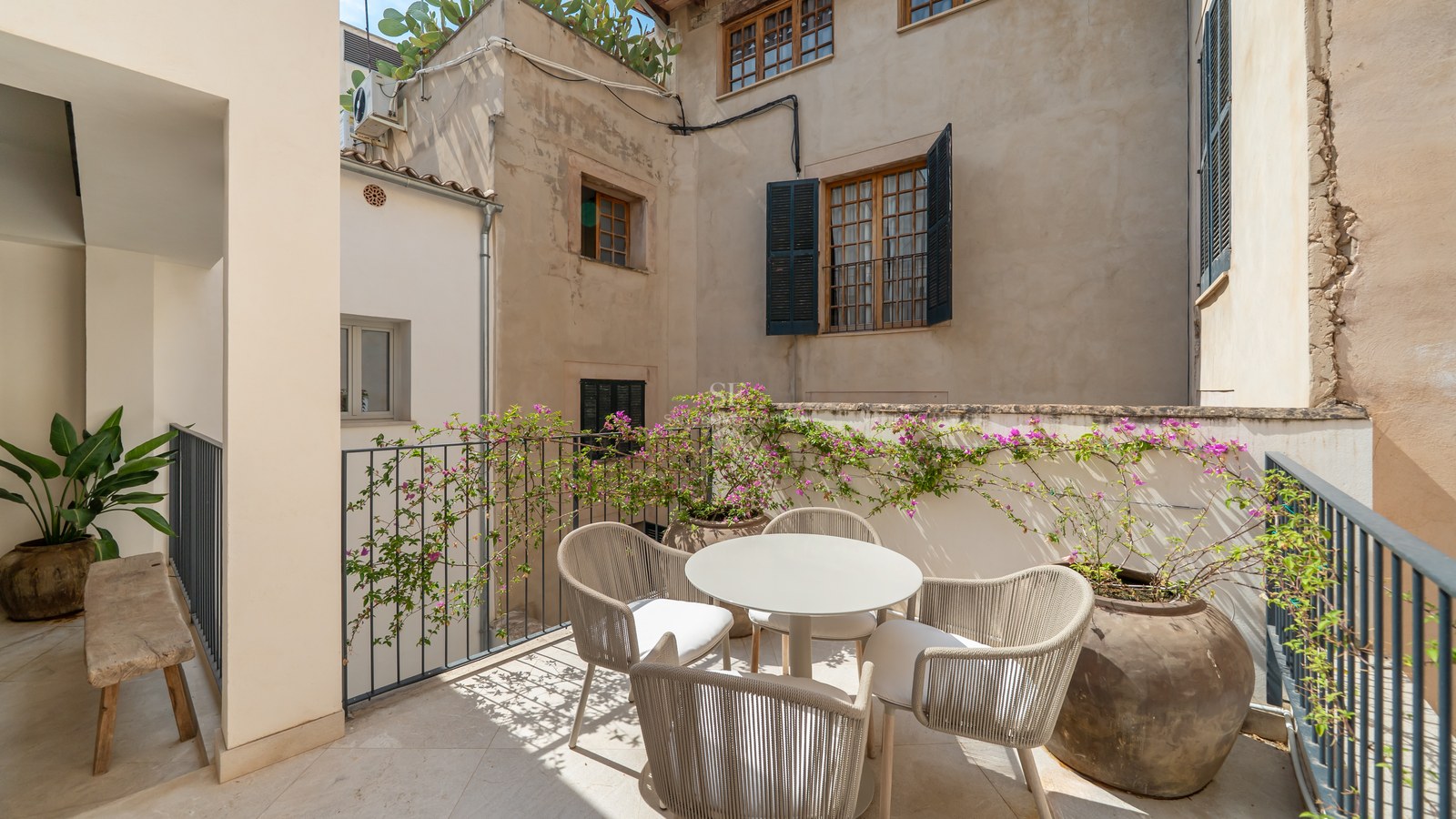 Sunlit terrace with modern outdoor dining set, surrounded by traditional stone walls, shutters, and flowering bougainvillea.