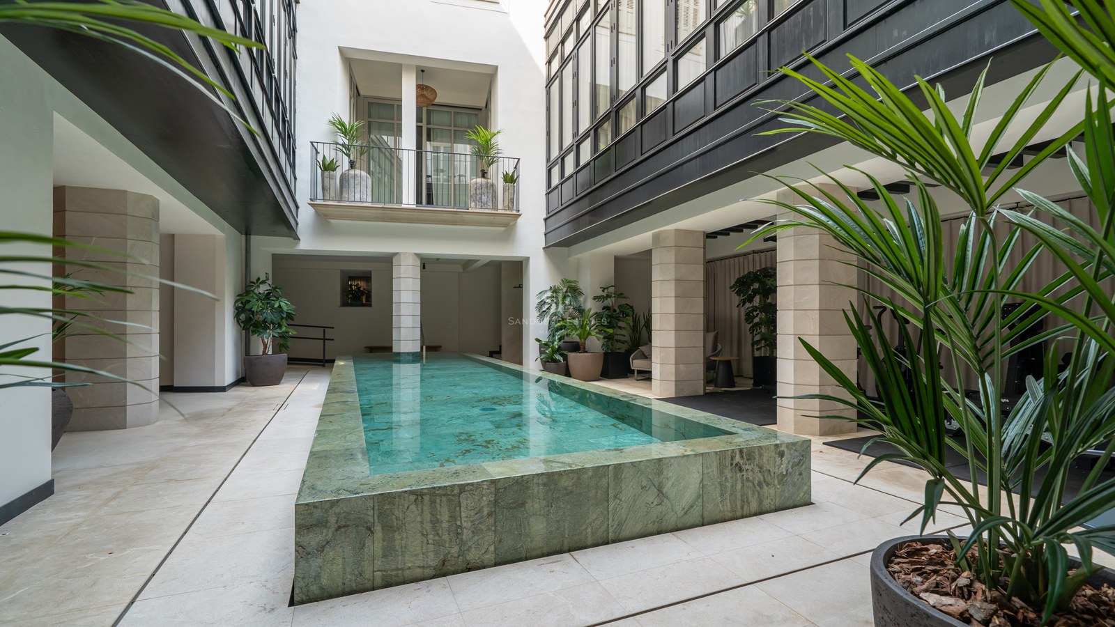 A rectangular raised lap pool made of green natural stone in a white-walled courtyard with columns and palms.