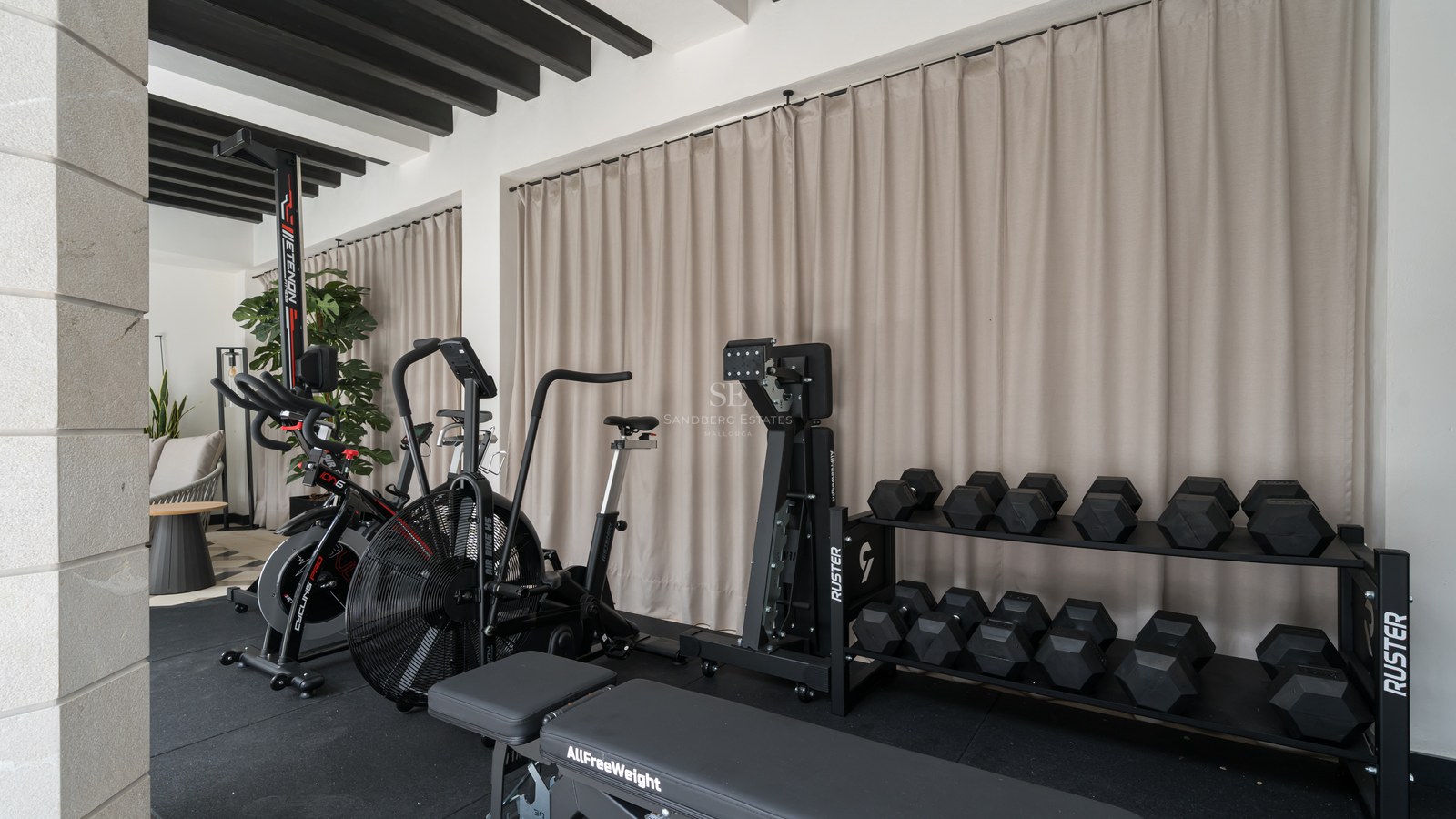 Modern home gym with stationary bikes, weight rack, and workout bench against beige curtains and black ceiling beams.