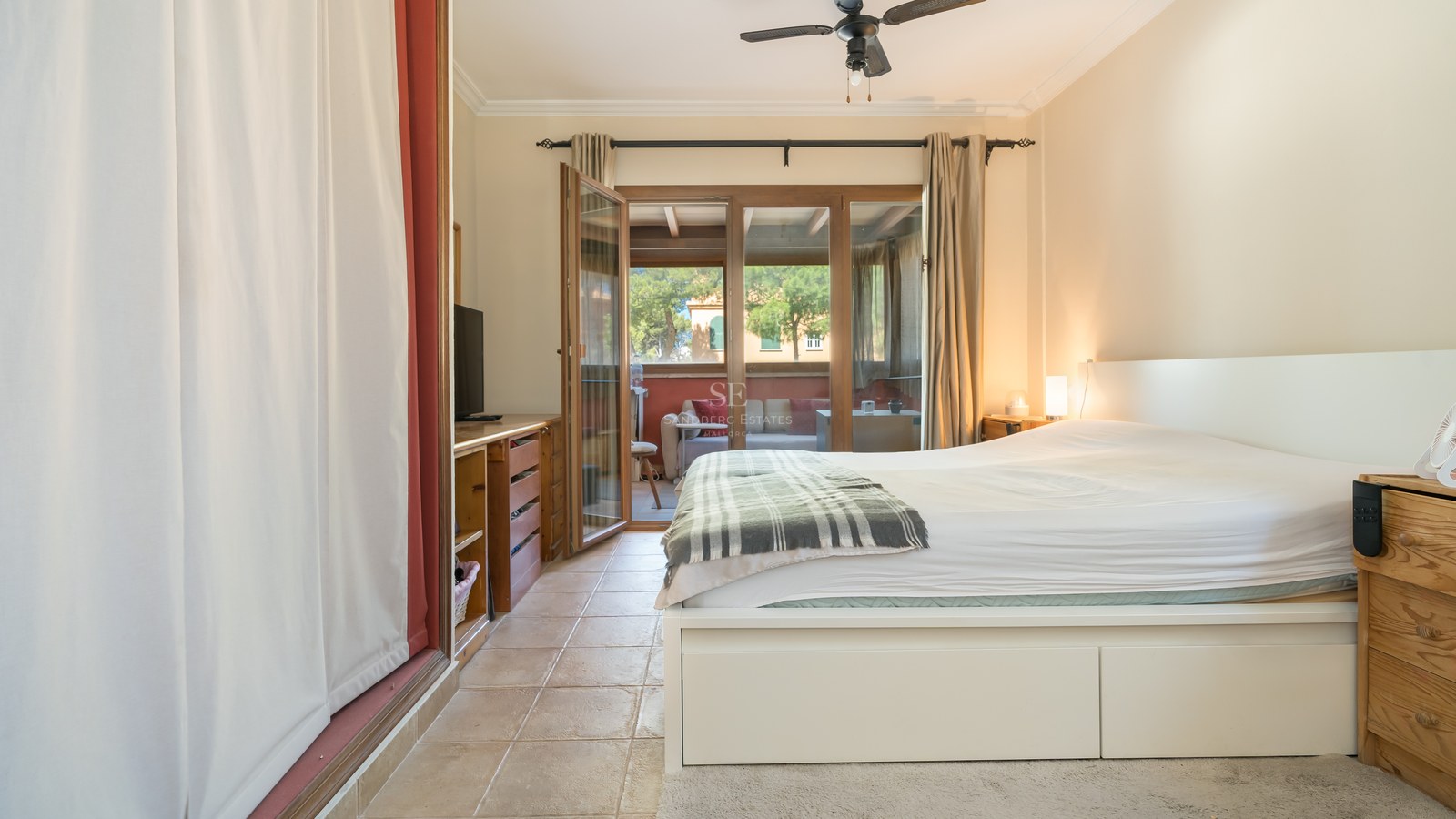 Dormitorio con suelos de terracota, cama blanca y puertas acristaladas que dan a una terraza amueblada.