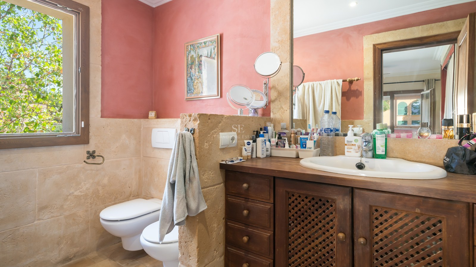 Cuarto de baño con paredes de piedra natural, mueble de madera oscura, inodoro y bidé, con ventana al jardín.