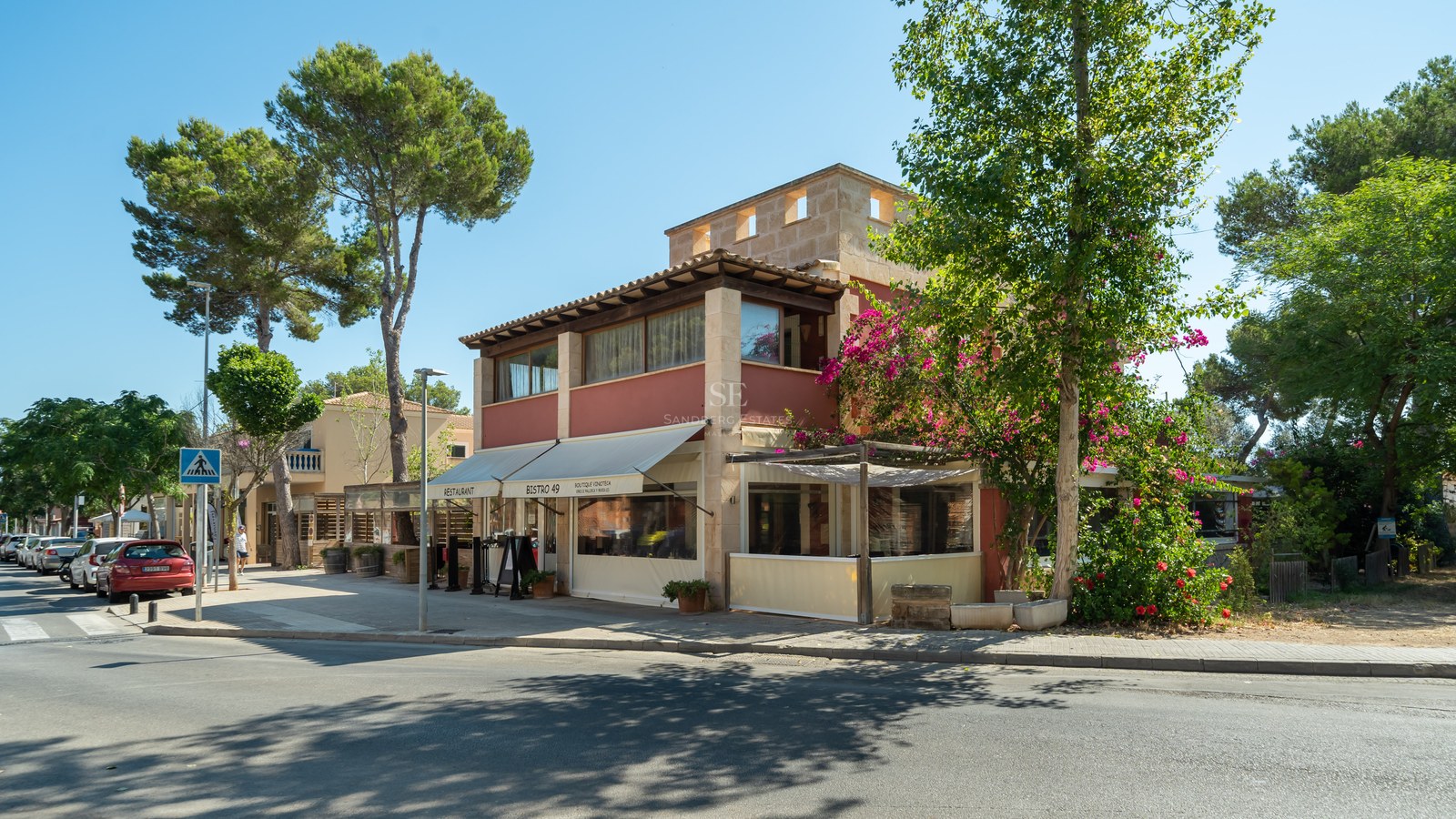 Edificio mediterráneo de dos plantas con un restaurante en la planta baja, rodeado de pinos y buganvillas.