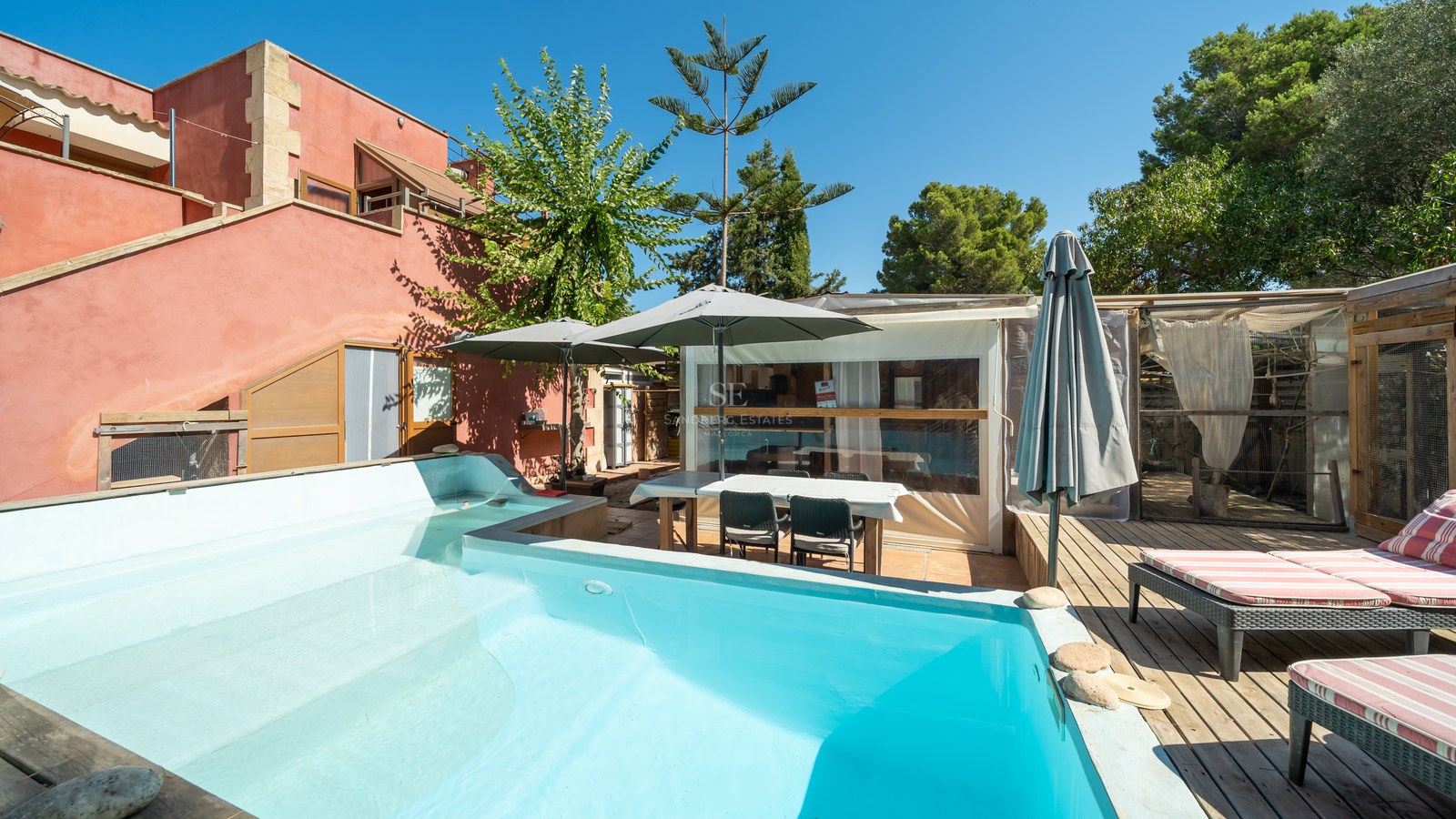 Piscina turquesa junto a terraza de madera con tumbonas y fachada de terracota bajo un cielo despejado.