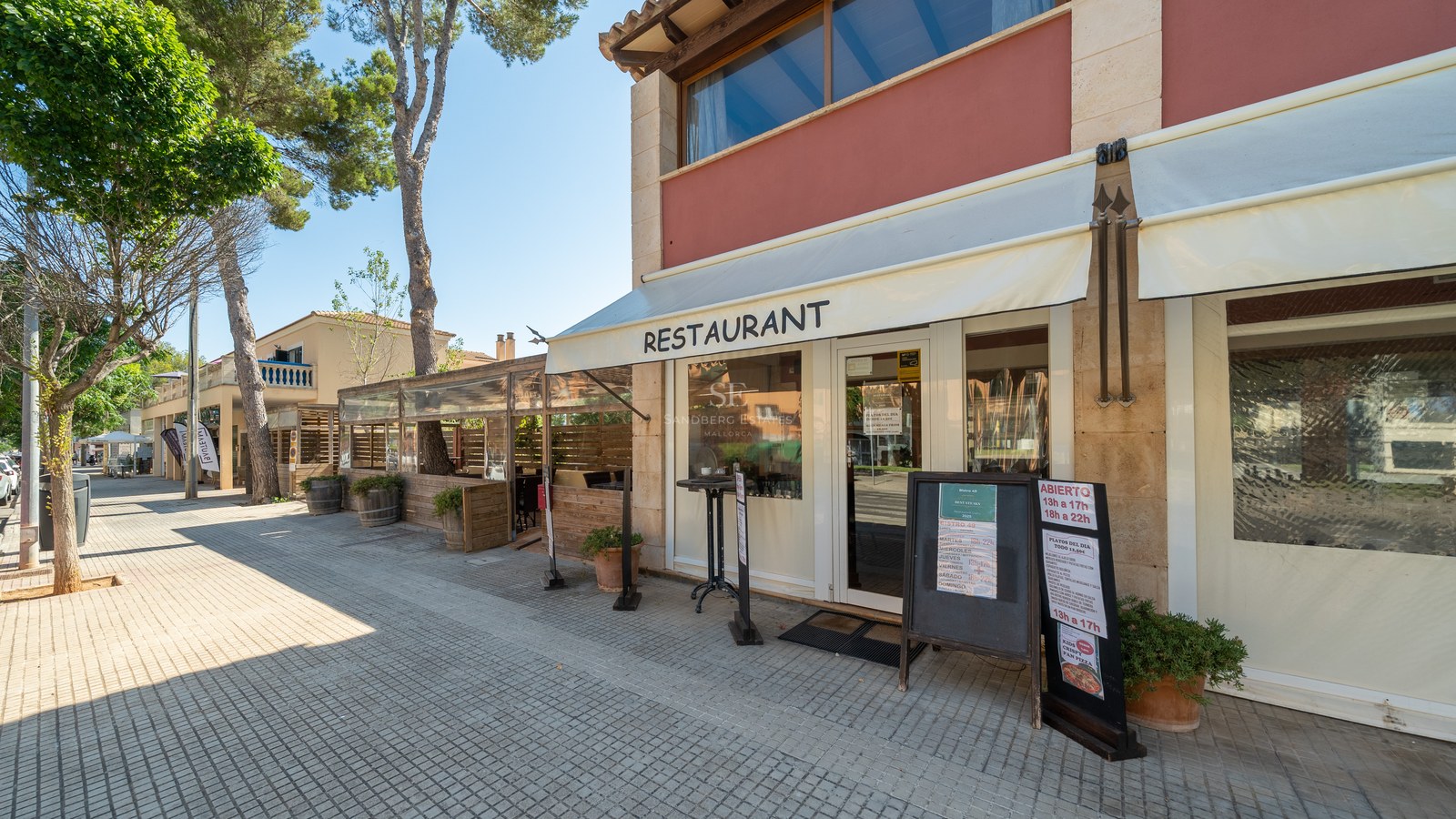 Vista exterior de un restaurante con paredes de piedra, toldos blancos y una acera arbolada bajo un cielo despejado.