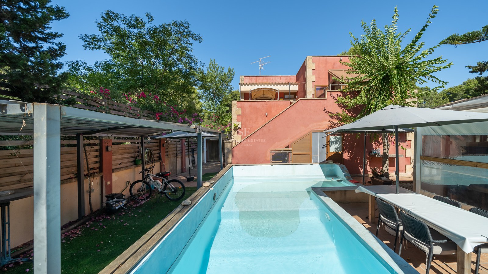 Piscina rectangular turquesa en un jardín privado con una casa color terracota y zona de comedor bajo sombrilla.