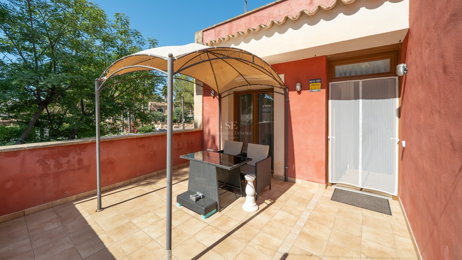 Terraza soleada con paredes de color terracota, suelo de baldosas y un cenador con mesa y sillas.