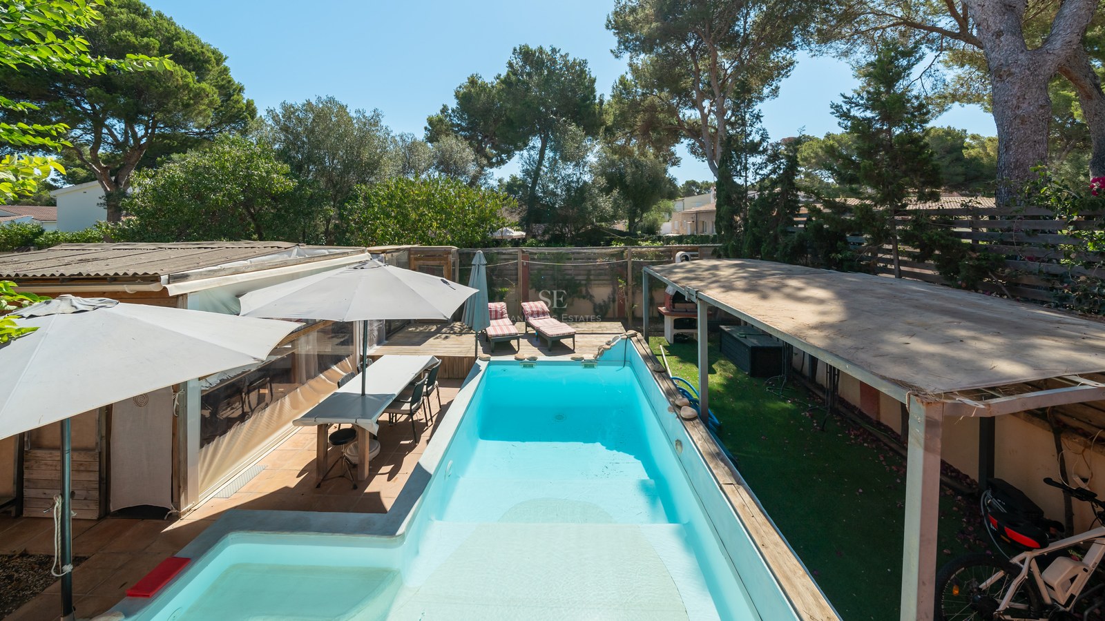 Piscina rectangular turquesa rodeada de tarima de madera, comedor exterior y árboles bajo un cielo azul despejado.
