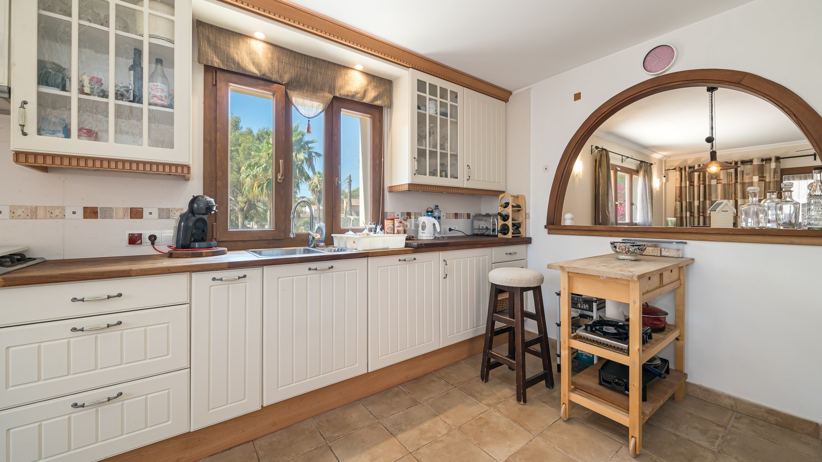 Cocina crema con encimeras de madera, suelo de terracota y ventana de arco hacia el comedor y jardín.