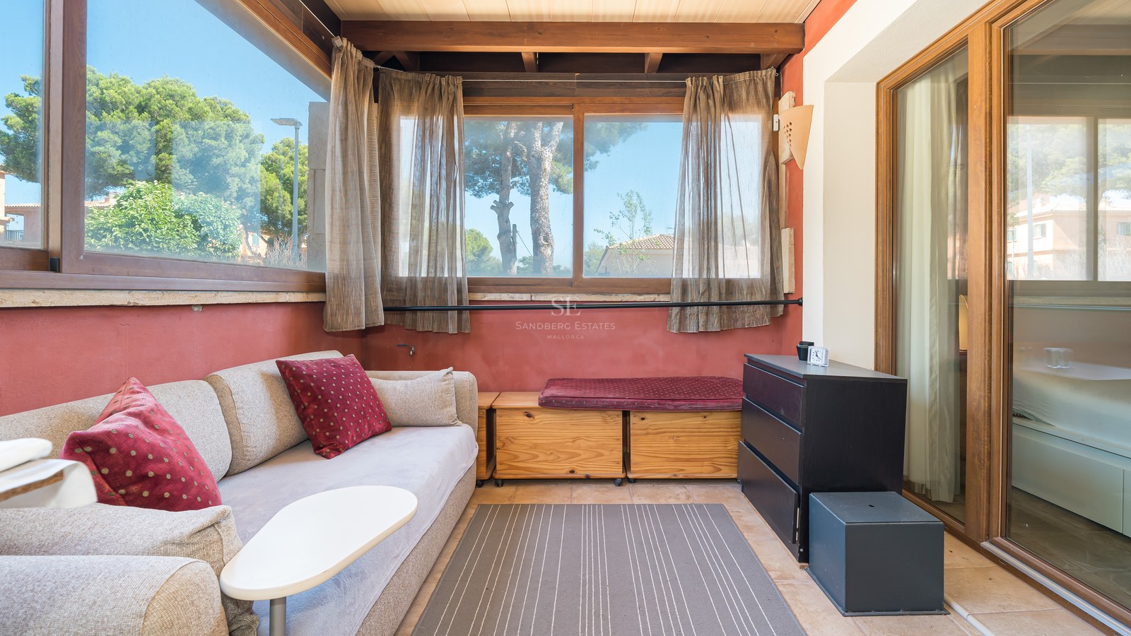 Sala de estar iluminada con sofá beige, vigas de madera, paredes de terracota y grandes ventanas con vistas al jardín.