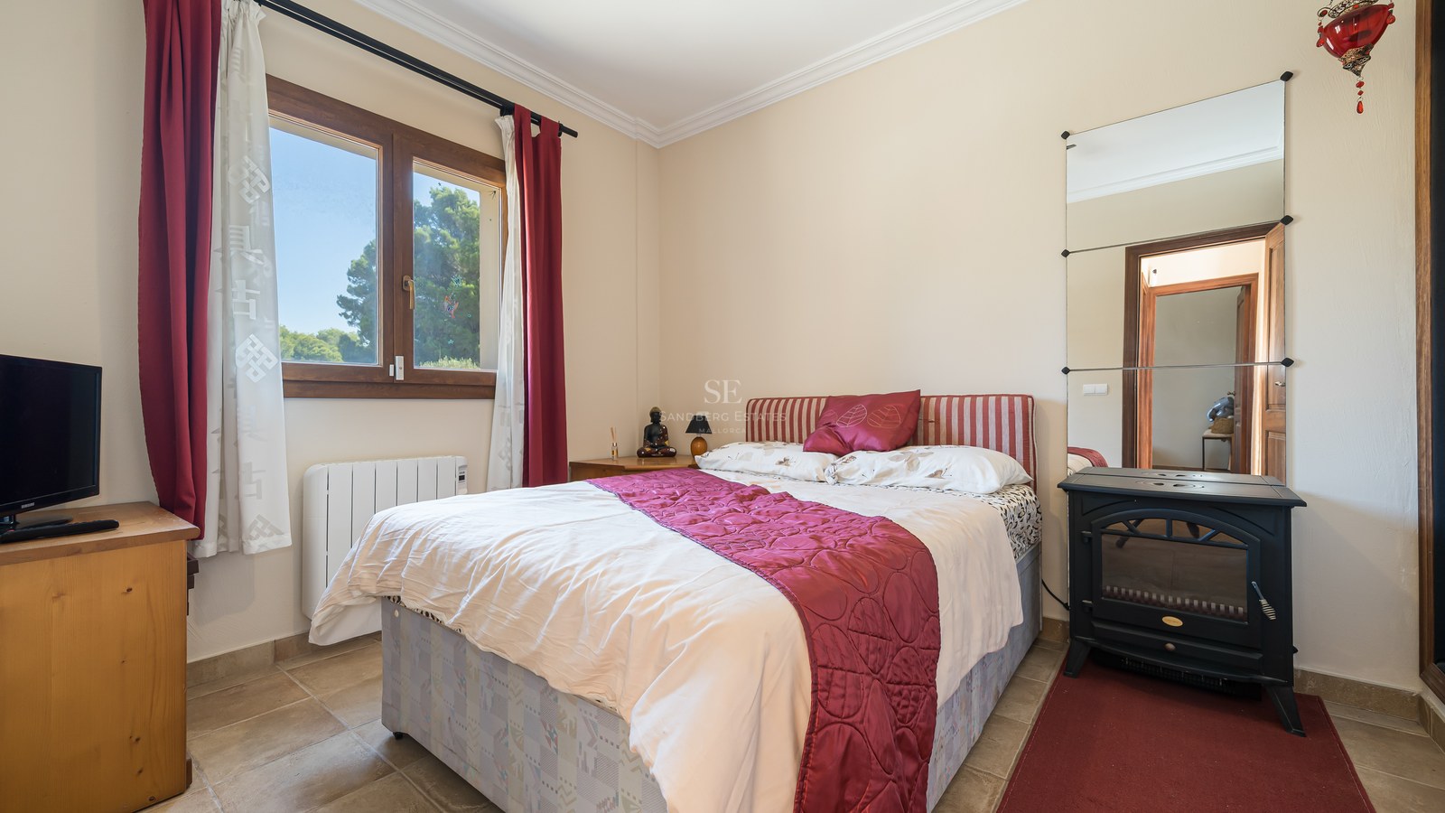 Dormitorio con cama doble, textiles granates, ventana de madera con vistas a pinos y estufa decorativa negra.