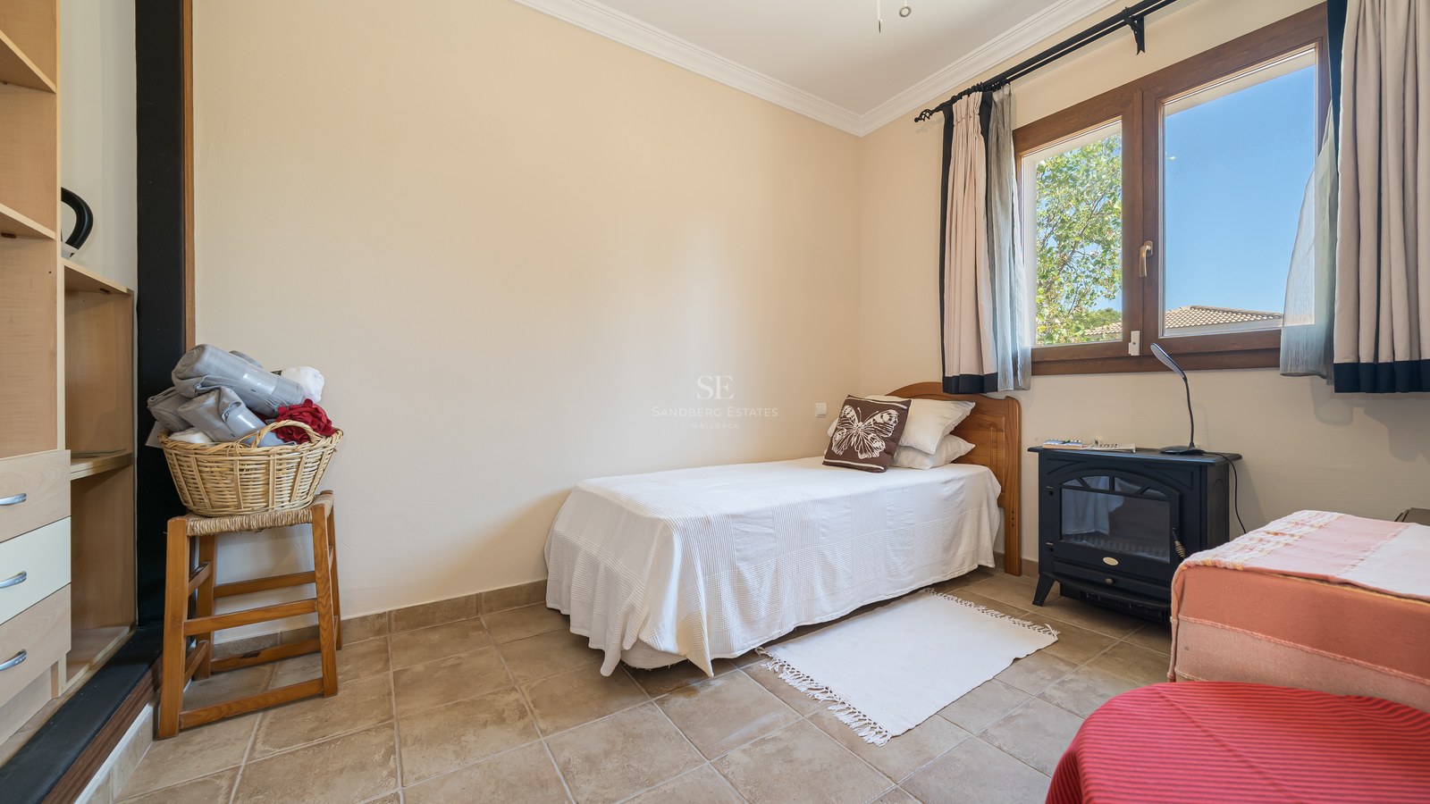 Pequeño dormitorio con cama individual, suelo de baldosas, ventana de madera y estufa decorativa.