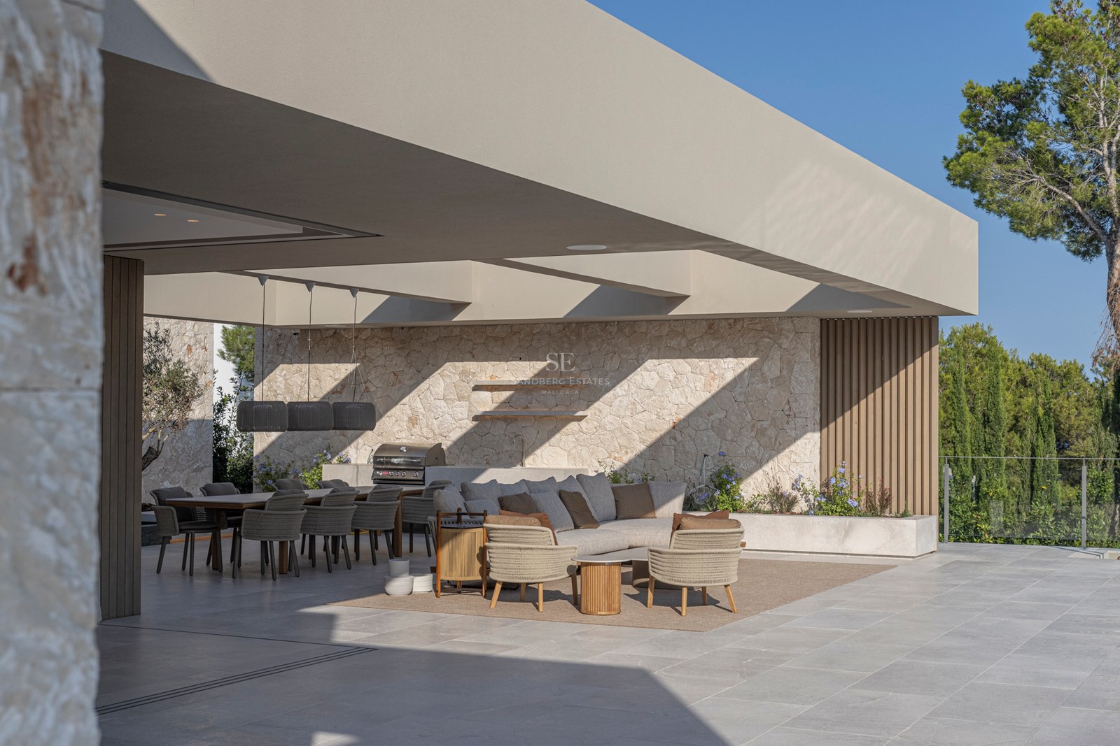 Terrasse de luxe avec salon gris, murs en pierre et espace repas sous un toit architectural moderne.