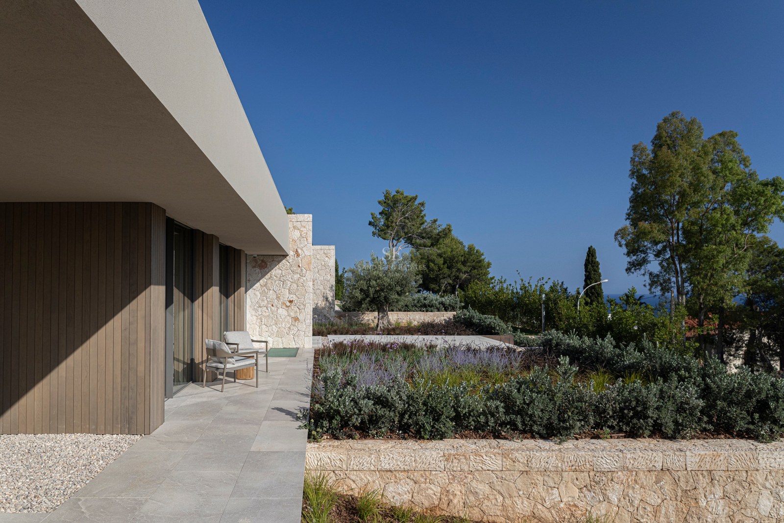 Terrasse de villa moderne avec fauteuils gris, murs en pierre naturelle et bardage en bois sous un auvent.