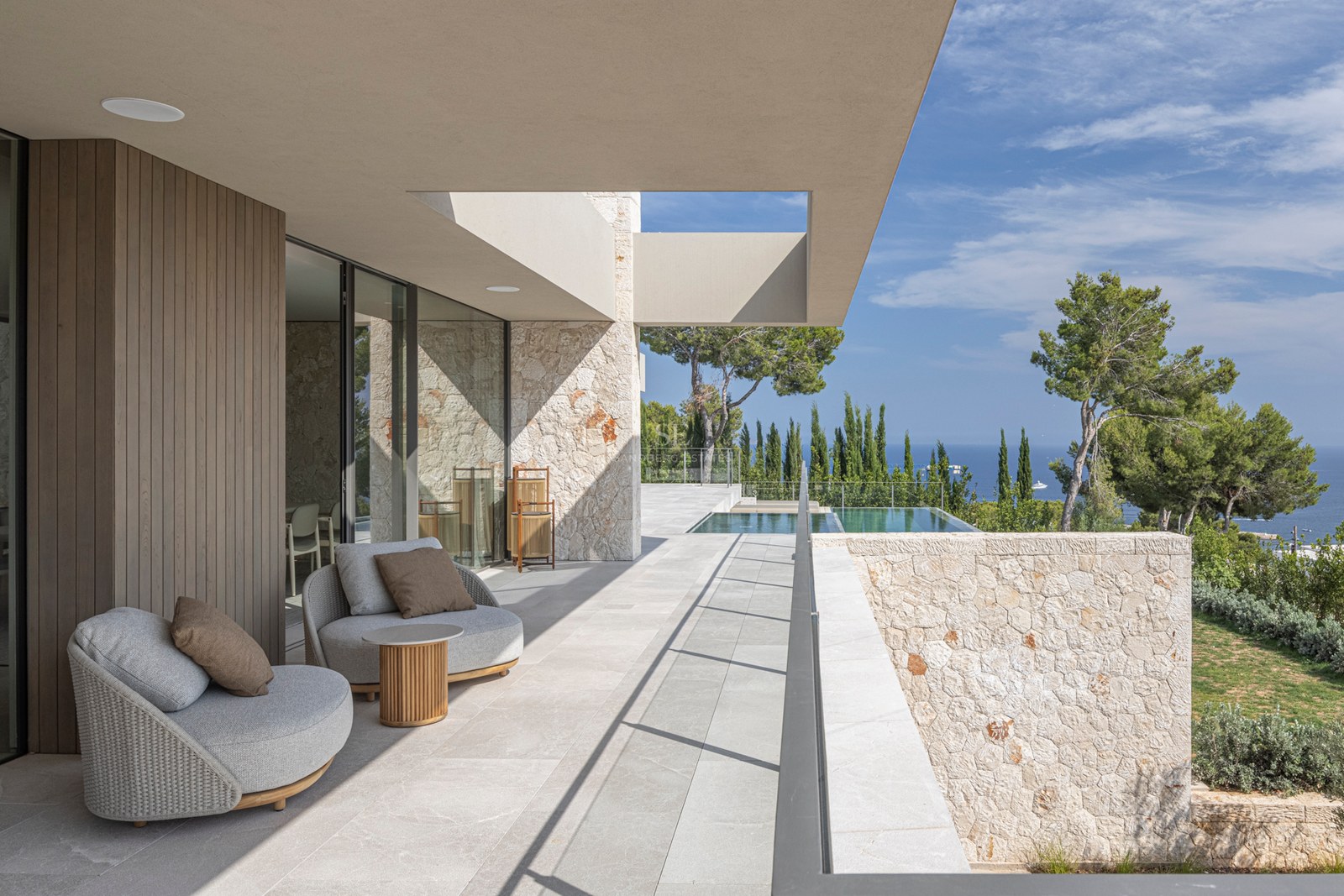 Terrasse de luxe avec fauteuils gris, murs en pierre et piscine à débordement surplombant la mer et les pins.