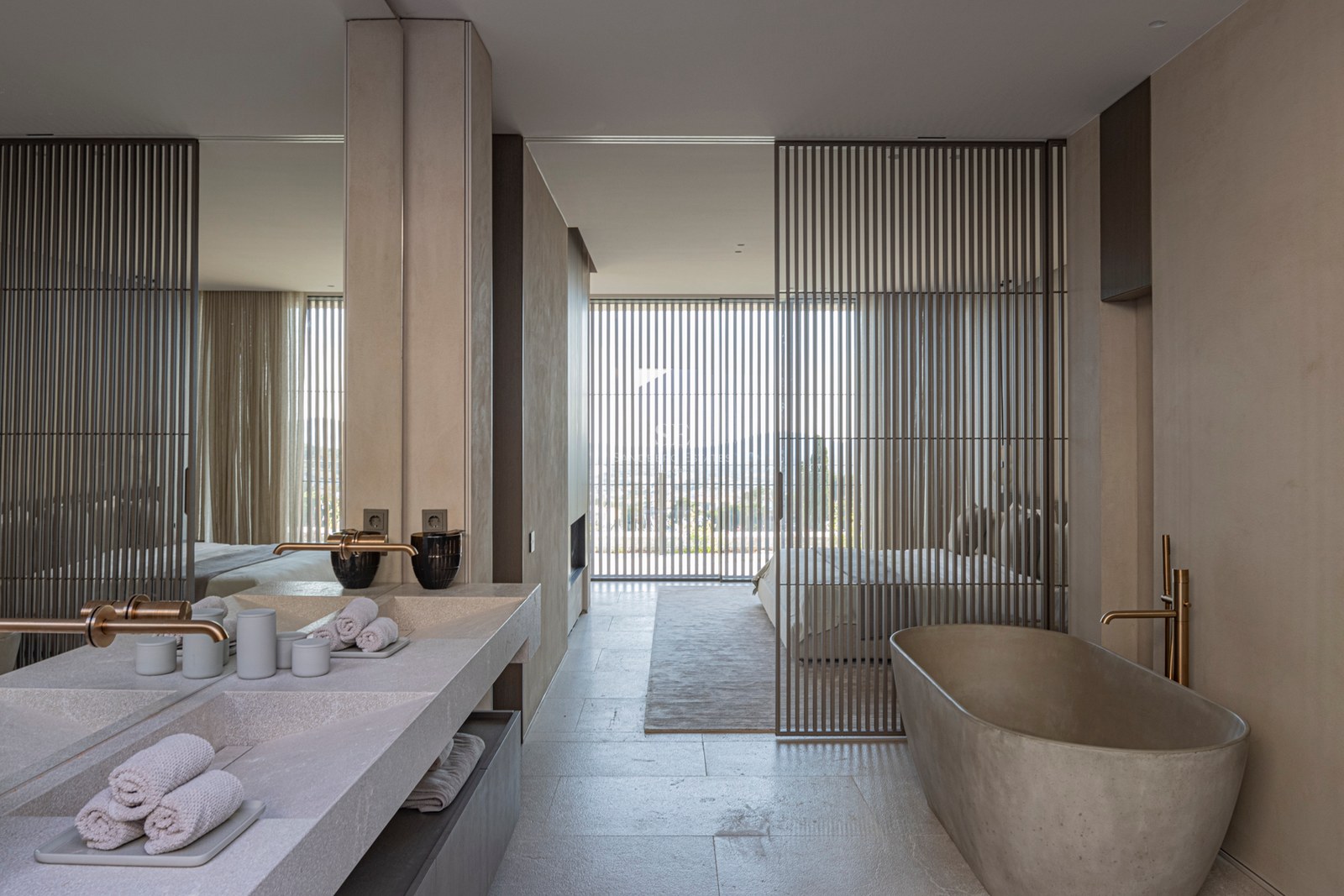 Salle de bain minimaliste avec baignoire en pierre, double vasque et laiton brossé, ouverte sur la chambre.