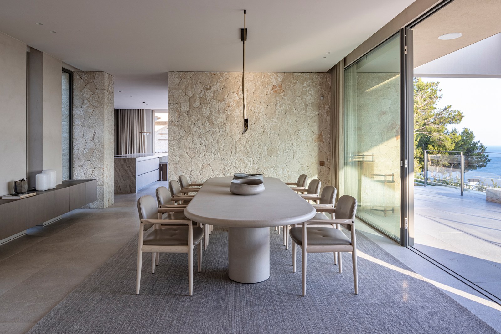 Salle à manger contemporaine avec table ovale en pierre, mur en pierre naturelle et grandes baies vitrées.