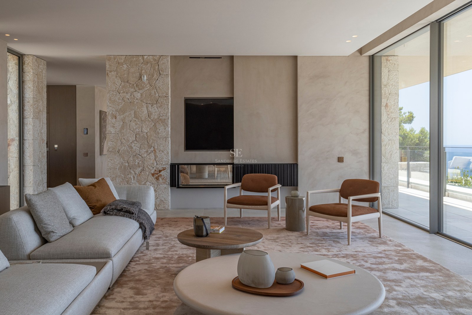Salon moderne avec murs en pierre, canapé beige, fauteuils terre cuite et grandes baies vitrées vue mer.