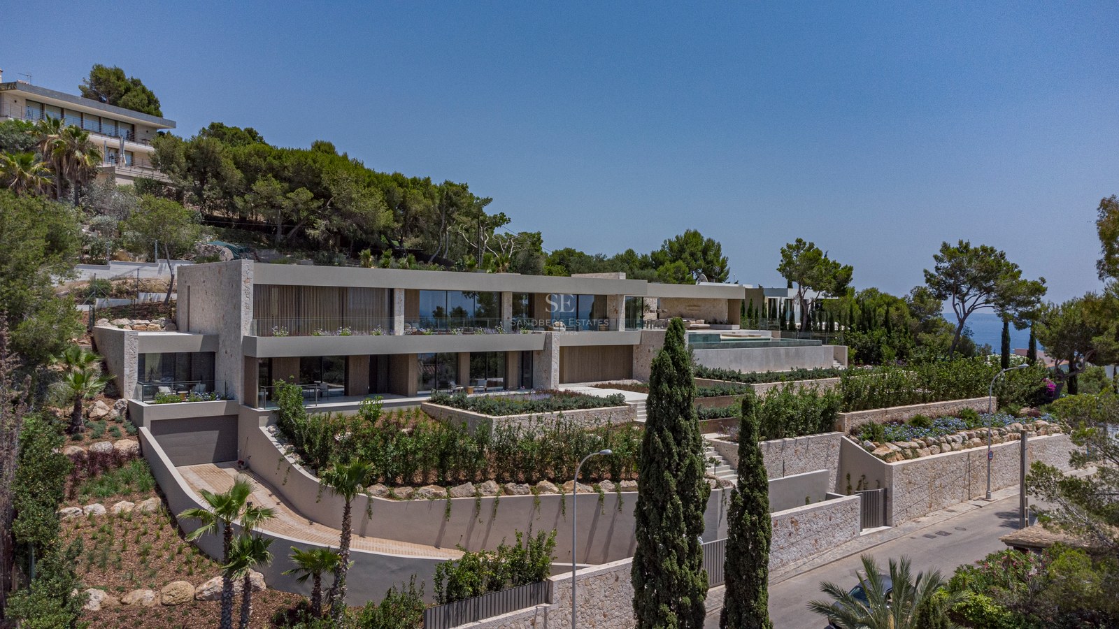 Villa moderne sur plusieurs niveaux sur une colline avec murs en pierre, garde-corps en verre et jardins méditerranéens.
