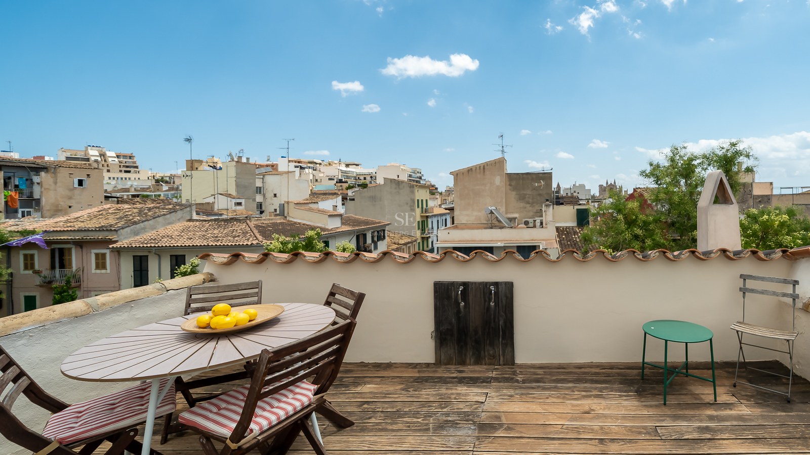 Terraza en la azotea con suelo de madera y zona de comedor con vistas a los tejados de una ciudad mediterránea.