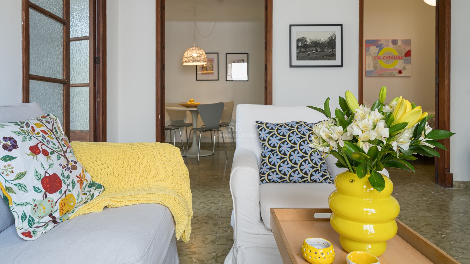 Salón luminoso con sofás blancos, acentos amarillos y vista al comedor a través de marcos de madera.