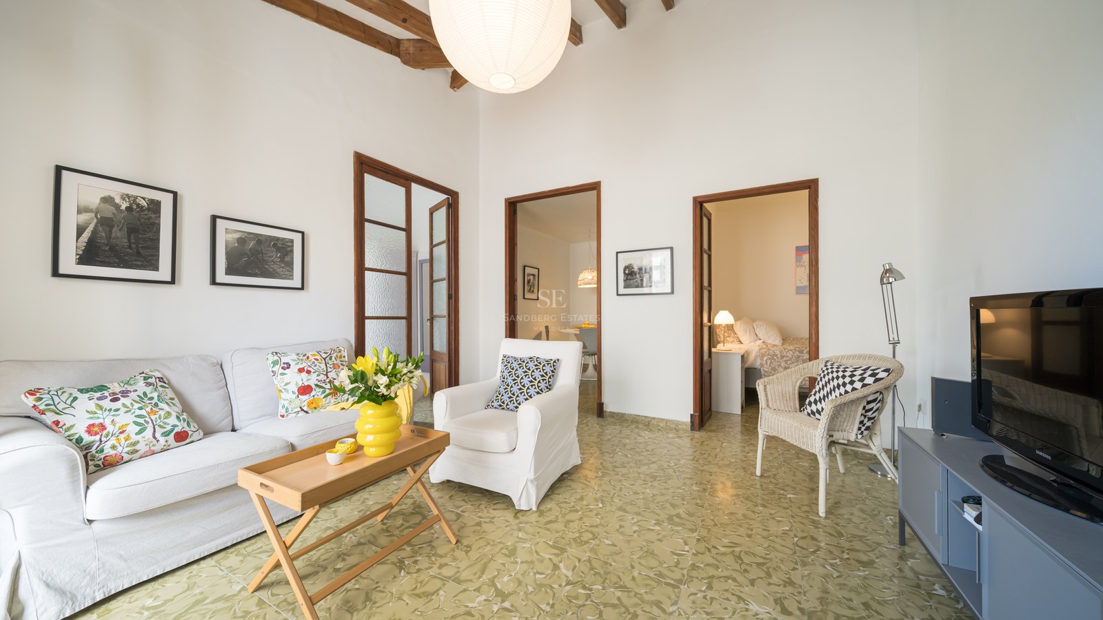 Salón luminoso con sofá blanco, vigas de madera rústicas, suelo de baldosas verdes estampadas y puertas a otras estancias.