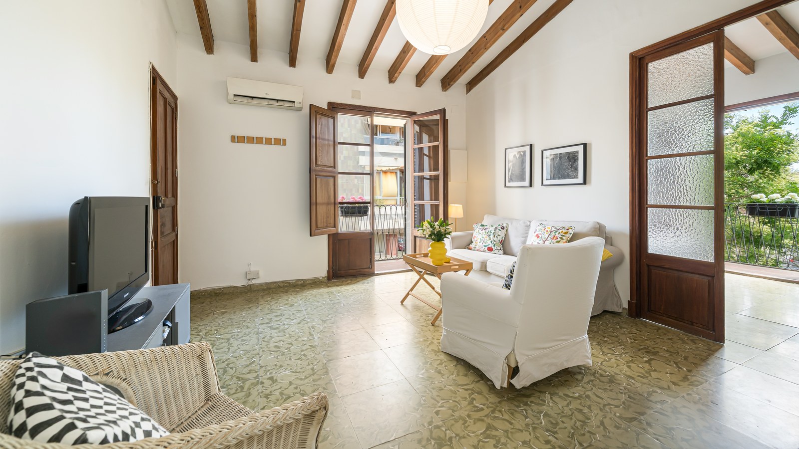 Salón con techos de vigas de madera, suelo de baldosa verde decorada, sofá blanco y puertas de balcón abiertas.