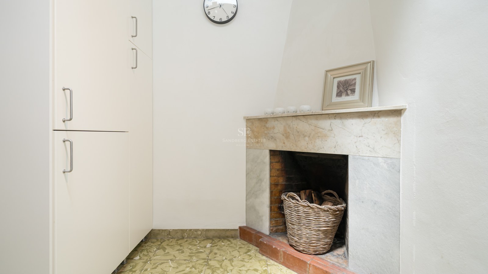 Rincón interior con chimenea de mármol, cesta de mimbre con leña y armario blanco alto.