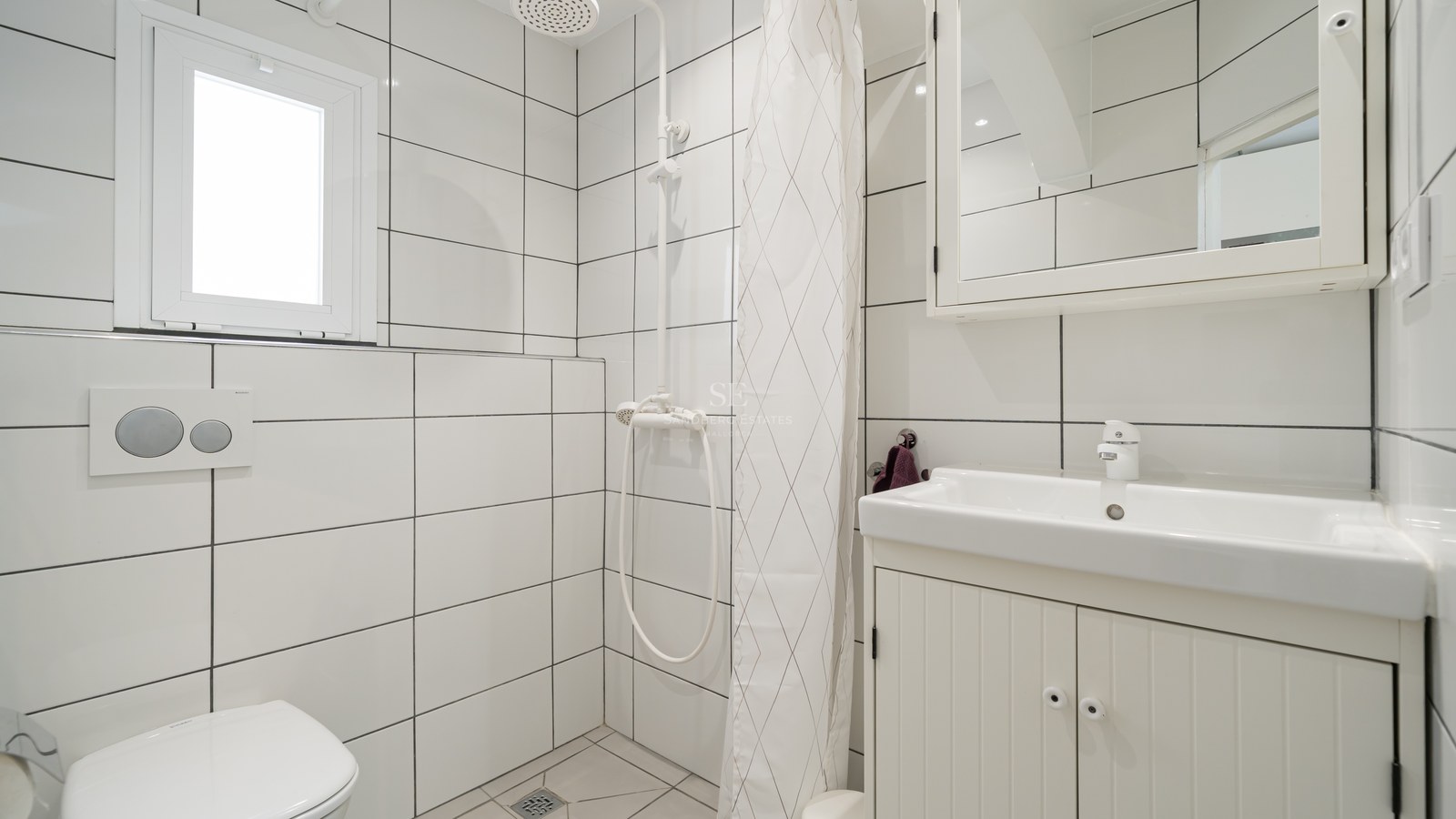 Baño alicatado en blanco con lavabo, inodoro y zona de ducha con cortina blanca.