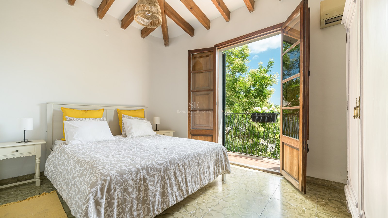 Un dormitorio luminoso con vigas de madera vista y puertas francesas que dan a un balcón soleado con árboles.