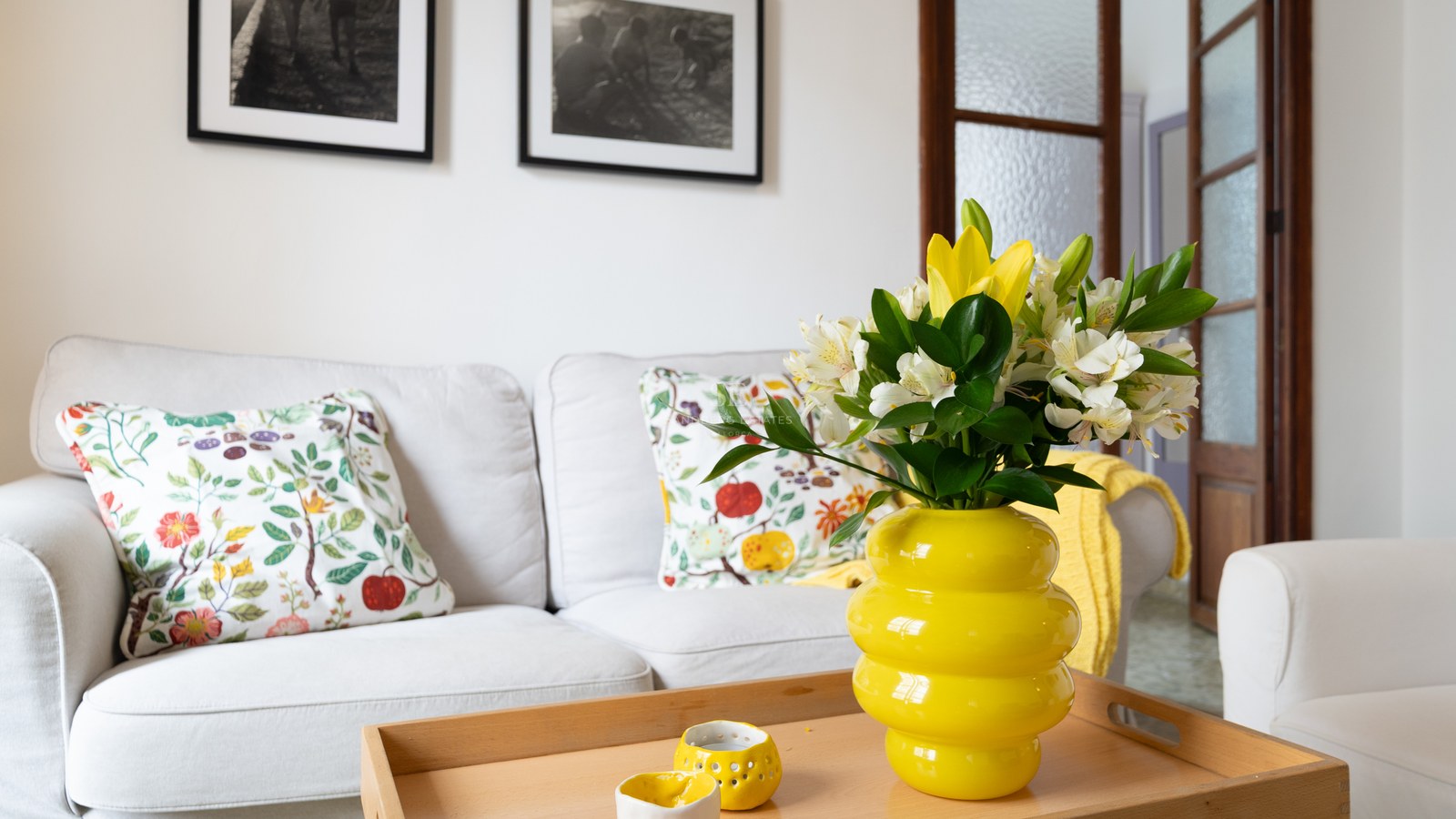 Sala de estar luminosa con sofá blanco, cojines florales y un jarrón amarillo vibrante sobre mesa de madera.