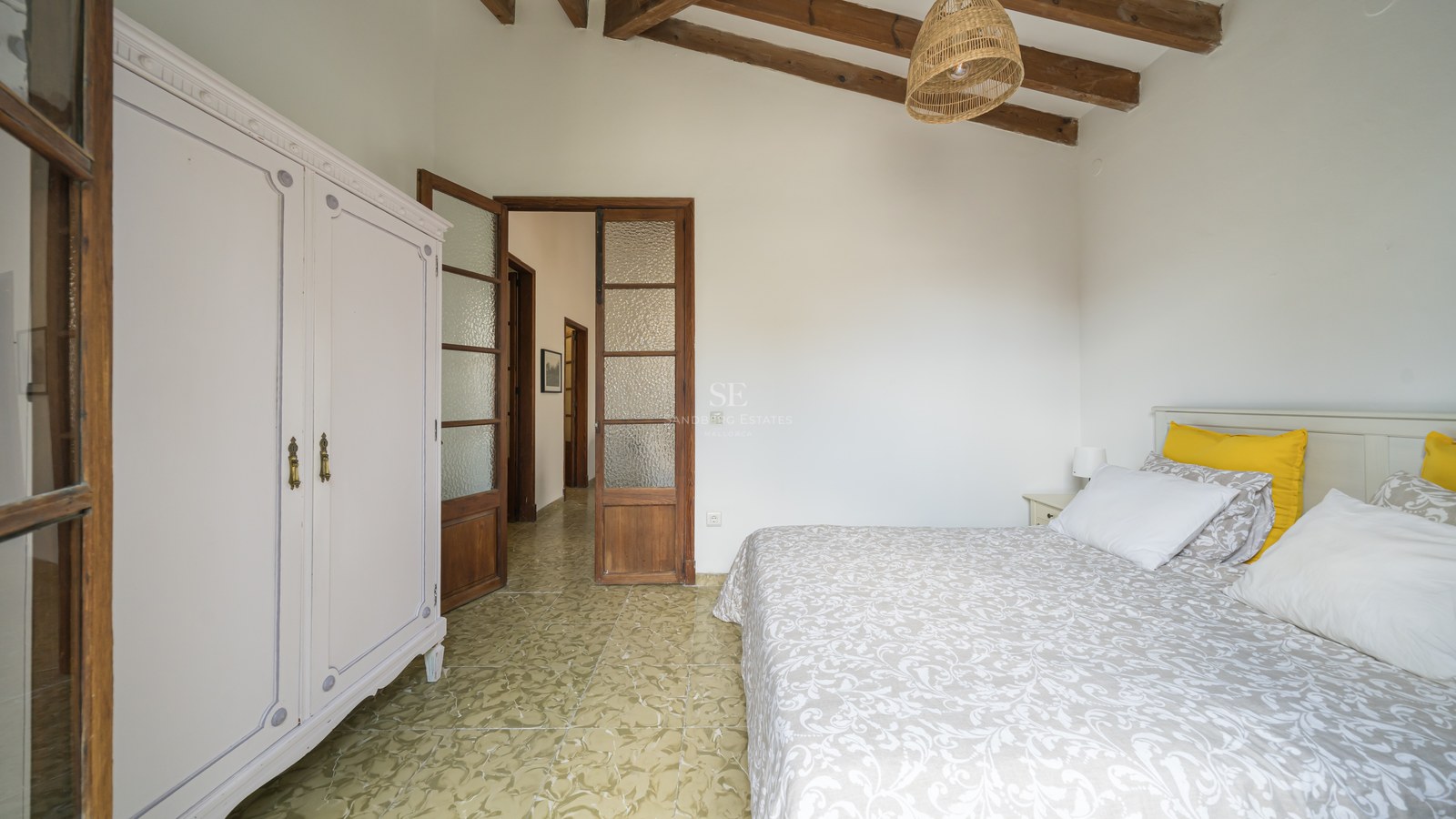 Dormitorio con vigas de madera en el techo, suelo de baldosas verdes estampadas, armario blanco y puertas rústicas.