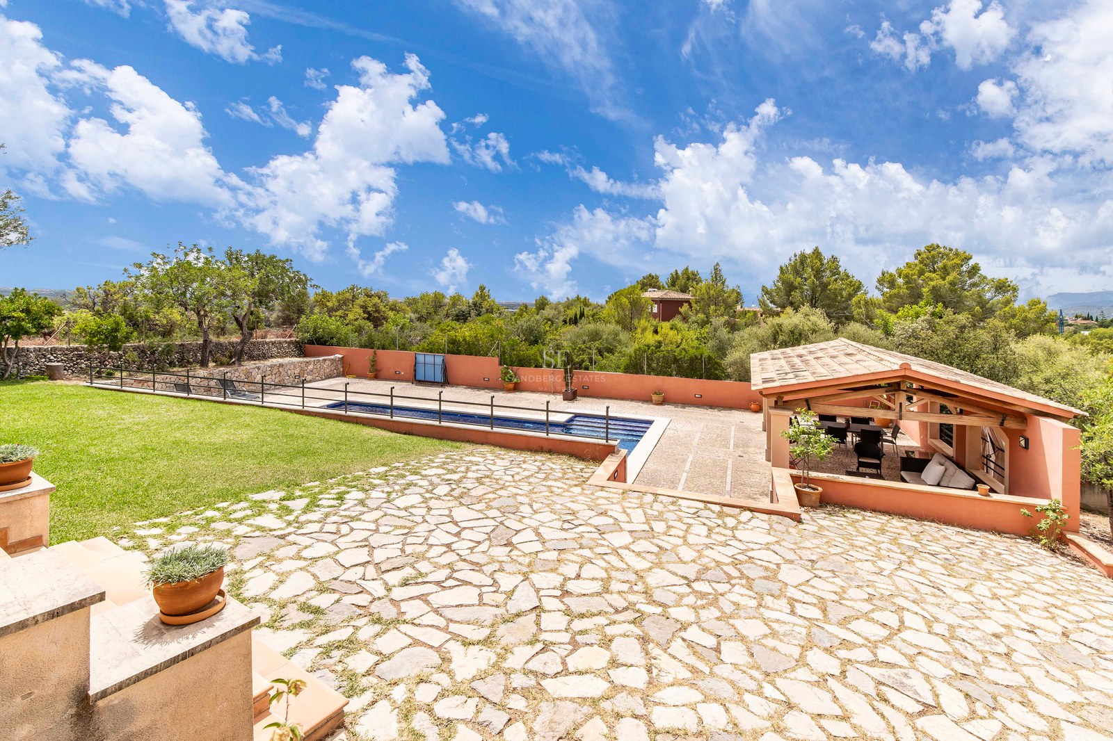Grand patio en pierre avec piscine, pelouse verte et salon couvert avec poutres en bois et toit en tuiles.
